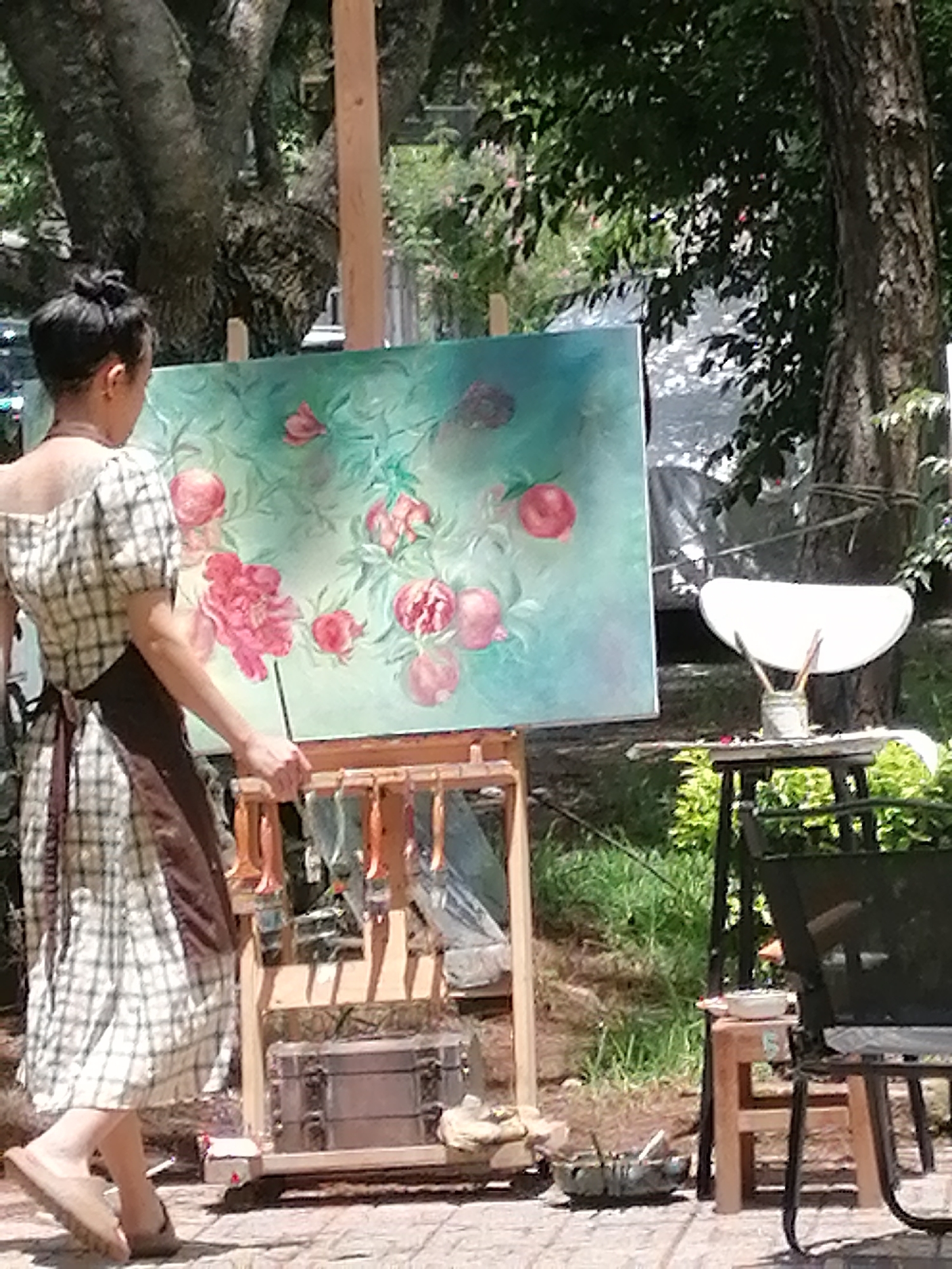 The image depicts an artist, viewed from the back, painting a large canvas displayed on an easel outdoors. The artist is wearing a checkered dress and an apron. The painting features a floral motif with pink flowers and green foliage against a teal background. In the foreground, a wooden easel holds the canvas and has various paintbrushes hanging from it. To the right, a small table holds a white palette and more brushes. A metal chest is visible beneath the easel. The setting appears to be a park or garden, with large trees and lush greenery in the background, suggesting a location like the Da Lat Botanical Gardens in Da Lat, Vietnam, known for its scenic outdoor spaces conducive to artistic activities. The ground is paved with light-colored tiles.