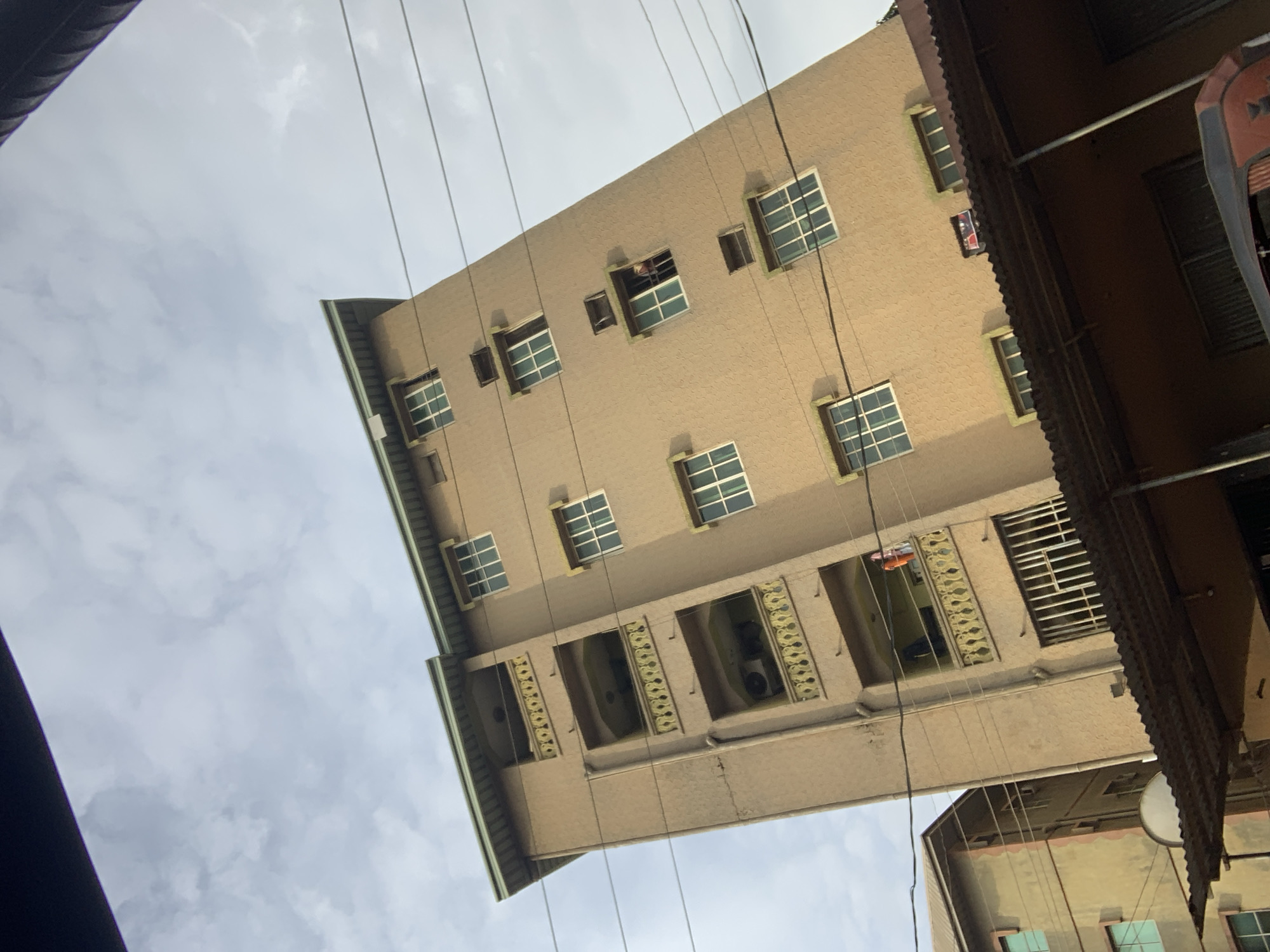 The image captures a low-angle view of a tall, multi-story residential building in Nkpor, Nigeria. The building is painted a light brown and features several balconies with decorative yellow railings on each floor. Windows are visible on the facade, some of which are open. In one of the open windows, a person wearing red clothing can be seen looking out. Another balcony has two figures visible, one of whom appears to be hanging laundry. Below the main building, a section with a corrugated metal roof is visible, supported by metal poles. A second, shorter building with similar window design is partially visible to the left. Overhead, power lines stretch across the frame, and the sky is overcast with clouds. The overall scene depicts a densely populated urban environment.