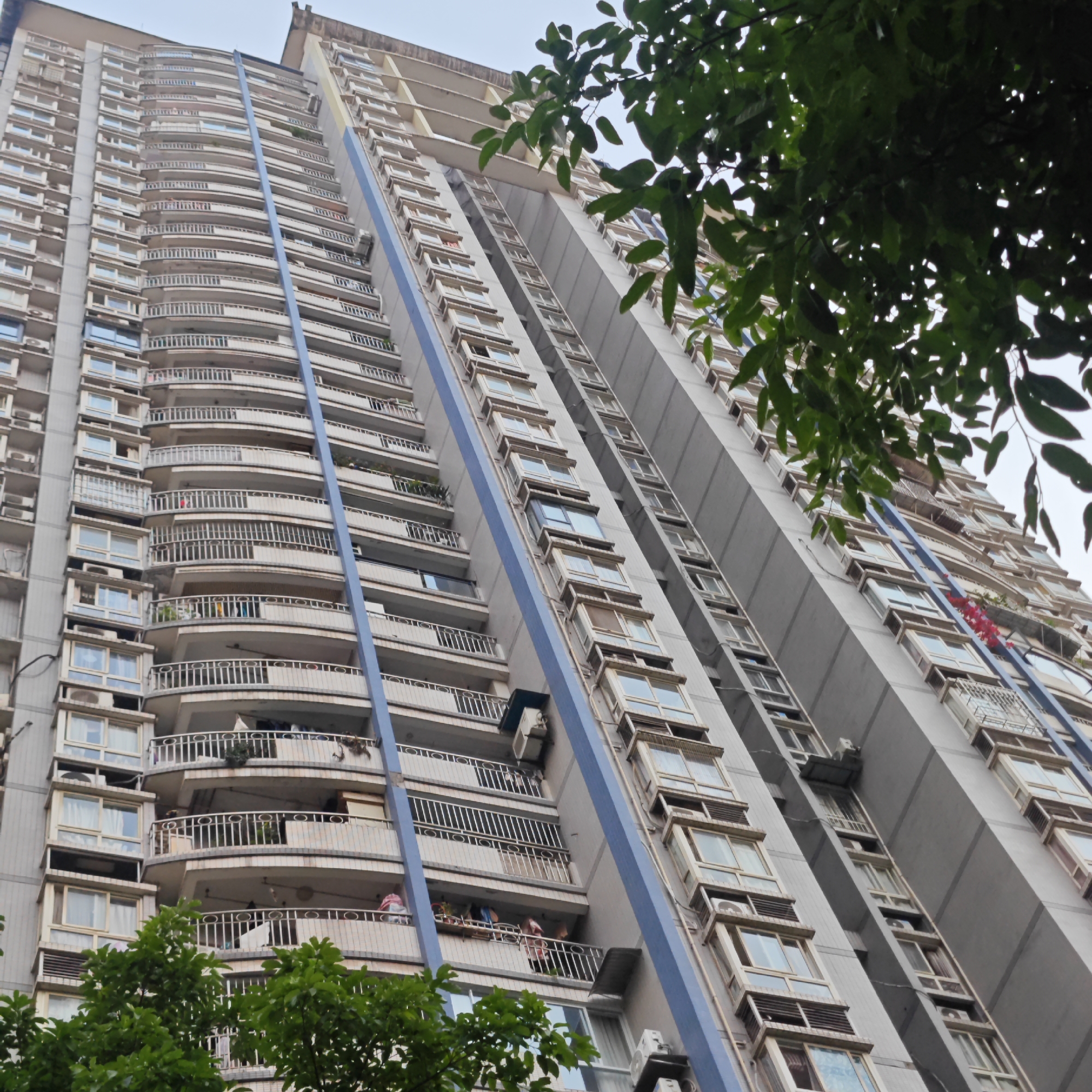 An upward-angle view of a high-rise apartment building in Yuzhong District, China. The building is tall with multiple stories, each featuring windows and balconies. The balconies have rounded railings. A blue stripe runs vertically along the building's facade. Green trees are visible in the foreground.