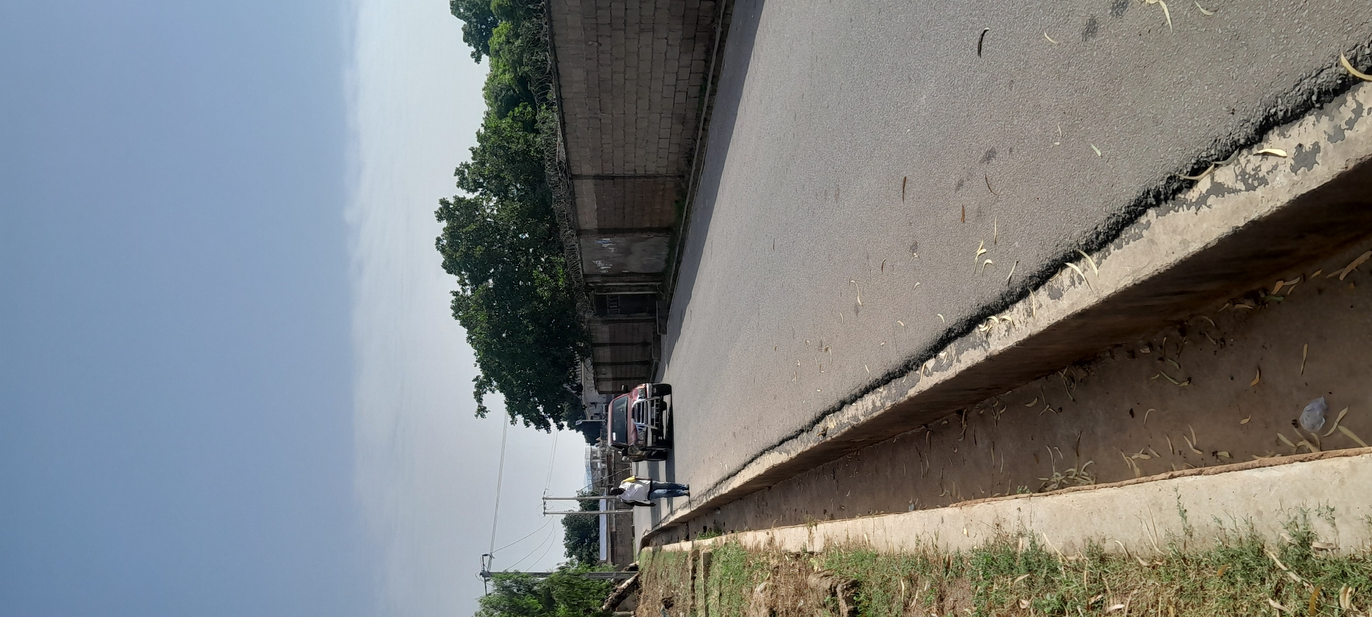 A mid-shot captures a person walking on the side of a paved road in Jalingo, Nigeria. The person, wearing a white short-sleeved shirt and blue jeans, is walking away from the camera, on the left side of the road. Ahead of the person, a red pickup truck is driving down the road. The road is lined with a concrete curb on the left, and a brick wall with barbed wire on top on the right. Trees are visible behind the wall. The sky is clear and blue. The image appears to have been taken in daylight.