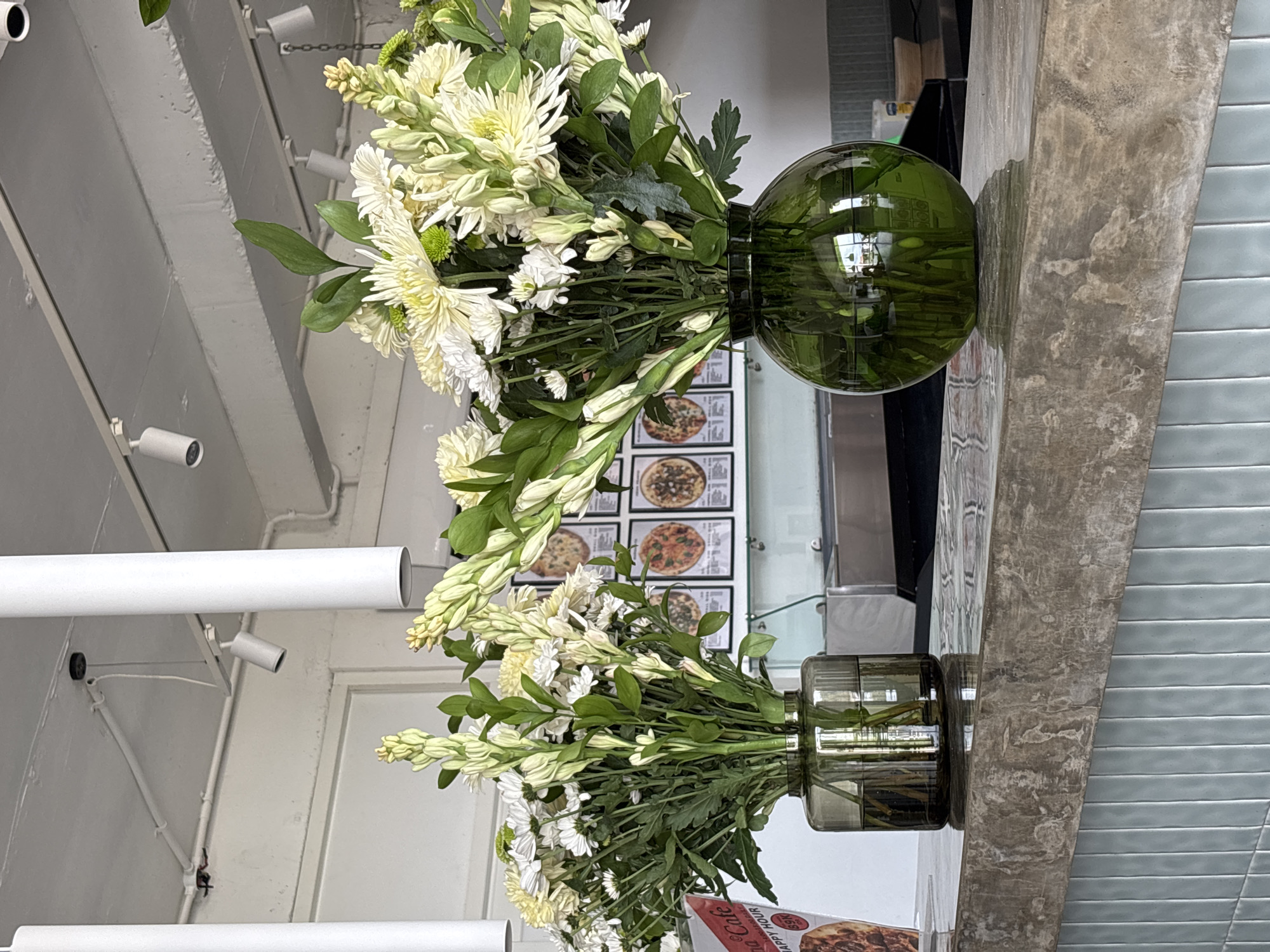 Two vases of white and green flowers are displayed on a counter. The vase on the left is a dark gray, cylindrical vase, and the vase on the right is a green, spherical vase. Behind the flowers, a wall features several framed pictures of pizzas and a menu board. The counter is made of concrete and is positioned in front of a light blue tiled wall. The overall setting appears to be a cafe or restaurant.