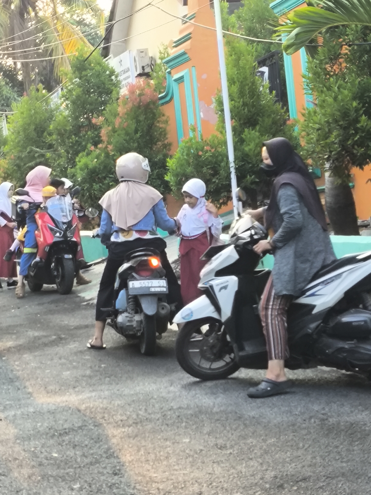 In Cilegon, Indonesia, a group of school-aged children in uniform and adults are gathered near parked motorcycles outside a building with an orange facade adorned with turquoise trim. Sunlight casts dappled shadows on the asphalt road. One child in a pink hijab and a red and blue uniform sits on a red motorcycle, holding onto a yellow helmet. Another child, wearing a white hijab and uniform, stands beside a person on a white motorcycle. This person, clad in a dark hijab, grey tunic, and striped trousers, is wearing dark clogs and appears to be preparing to ride. In the foreground, another motorcycle with a license plate reading "A 5577 SJ" is parked. The background features lush green trees and the side of a building.