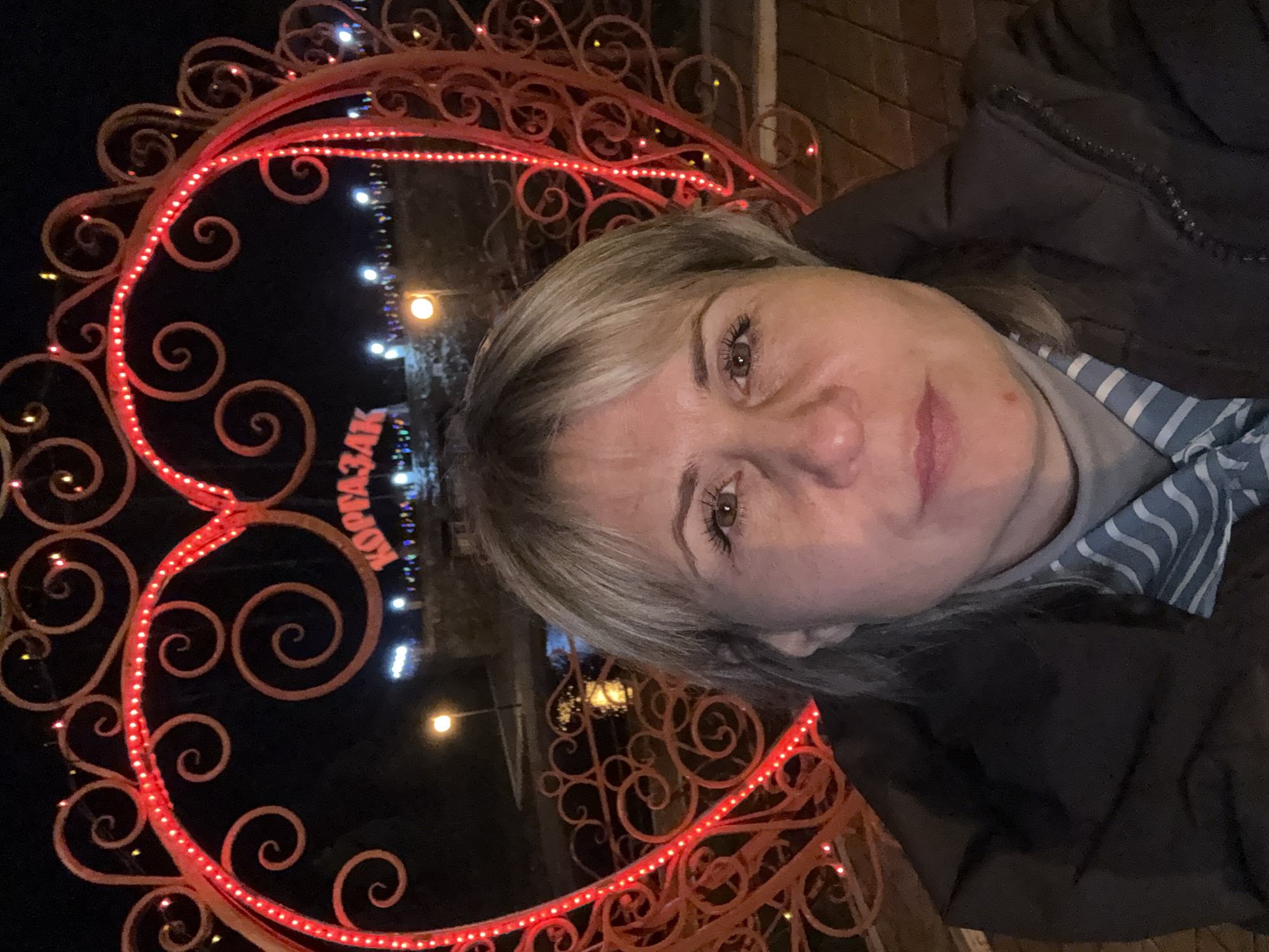 A woman with light brown hair stands outdoors at night in front of a decorative wrought-iron heart-shaped structure illuminated with red lights. The sign behind the structure appears to read "KOMSOMOLSK". The woman is wearing a dark jacket over a light blue and white striped shirt. The background includes other decorative lights. The location is likely Komsomolsk, Russia, based on the sign.