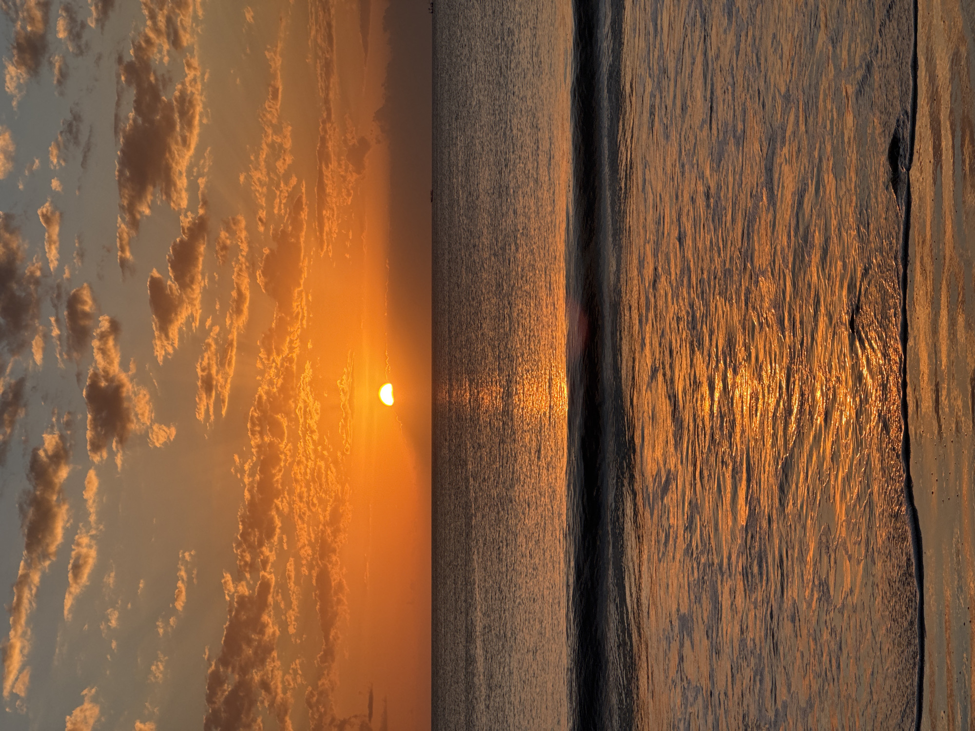 The image captures a serene ocean sunset at Kuta Selatan, Indonesia. The sky is a vibrant gradient of orange and yellow, adorned with scattered, fluffy clouds that catch the warm hues of the setting sun. A portion of the sun is visible just above the horizon, casting a shimmering golden path across the calm sea. Gentle waves lap at the shore, their crests reflecting the intense colors of the sunset. In the distance, two small vessels can be faintly seen on the water, adding a touch of scale to the vast expanse of the ocean. The overall atmosphere is tranquil and picturesque, embodying the beauty of a tropical sunset.