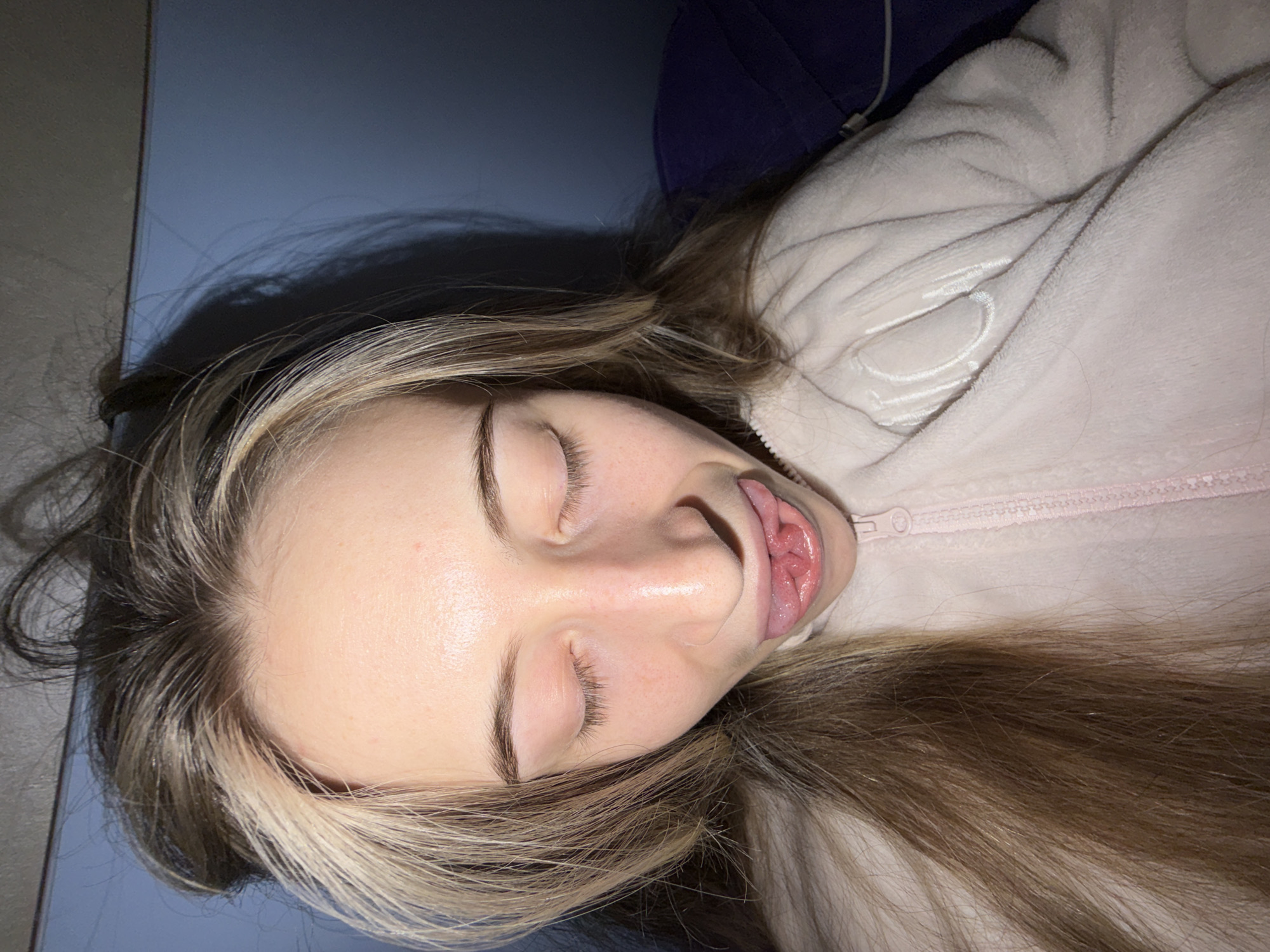 A young woman is lying on her side, her eyes closed and her tongue slightly protruding from her mouth. She has long brown and blonde hair, and is wearing a light pink fuzzy jacket. The surface she is lying on appears to be blue. The location is not discernible from the image and cannot be determined to be Plovdiv, Bulgaria.