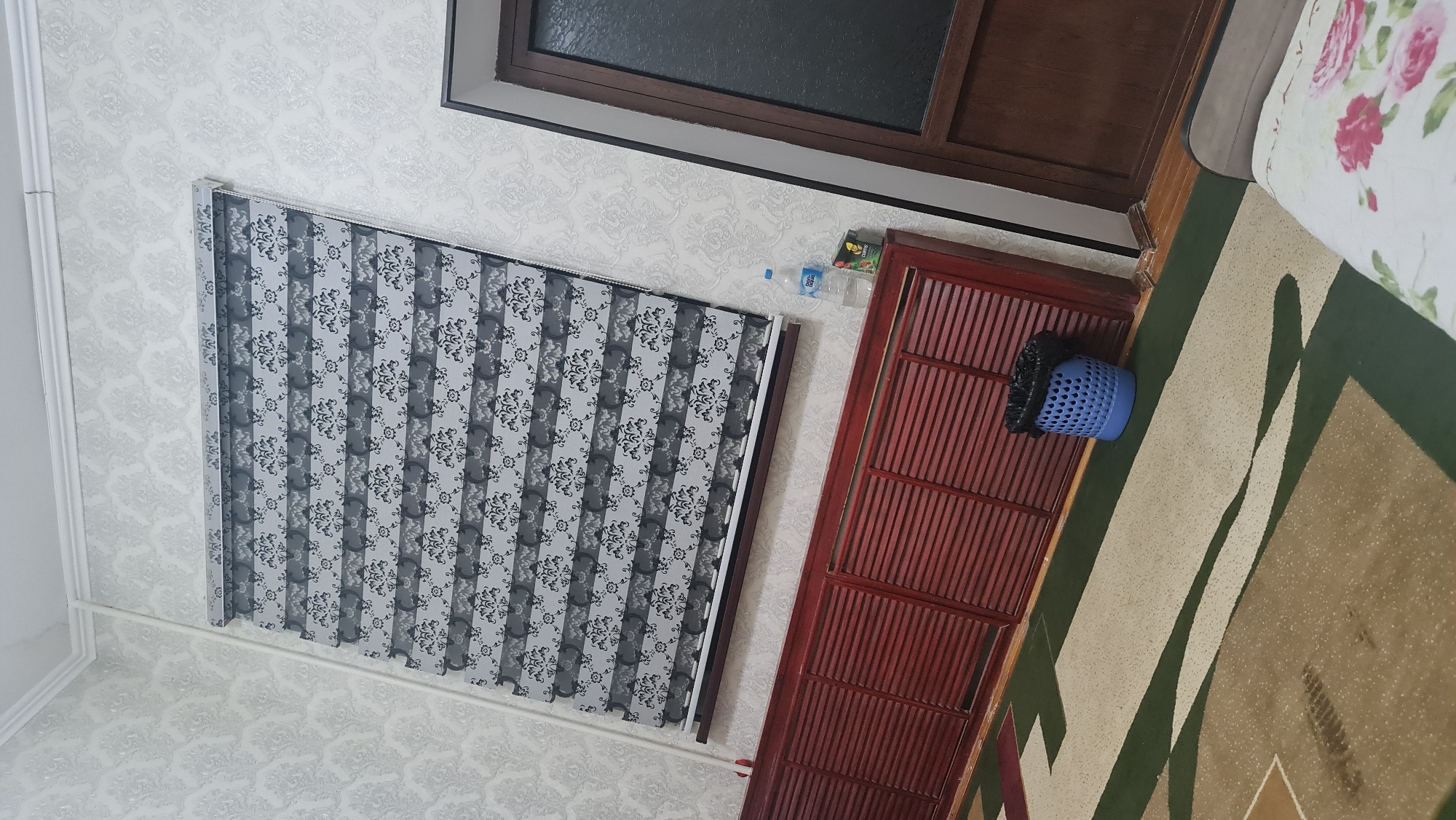A domestic interior scene, likely a bedroom or living area, is depicted from a slightly elevated perspective. The room features a patterned wallpaper with a damask motif in light grey and white. A horizontal blind with alternating opaque and sheer stripes, also featuring a dark floral pattern, is drawn across a window. Below the blind, a long, dark red wooden cabinet with louvered doors spans the lower portion of the wall. To the right of the cabinet, a blue plastic trash can with a black liner is visible. Further to the right, a portion of a wooden door with frosted glass is present. In the foreground, a carpet with a geometric design in cream, green, and brown tones covers the floor. A bottle of water and a small packaged item are on top of the cabinet. The scene is static and appears to be a still photograph. The location is identified as Karshi City, Uzbekistan.