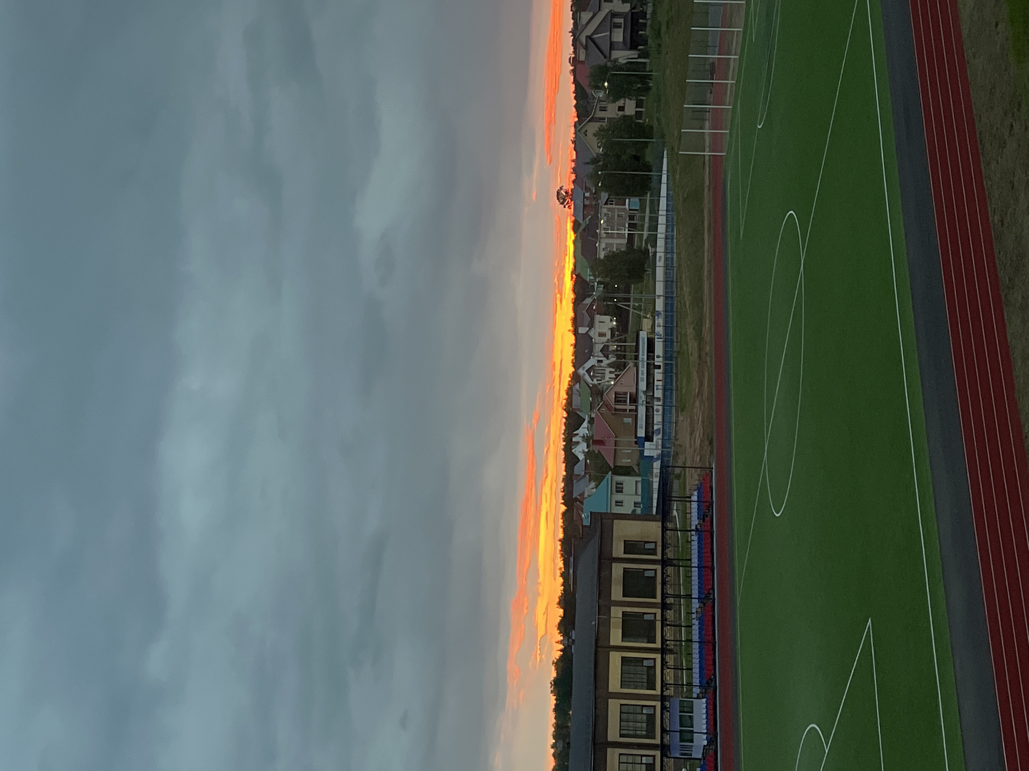The image shows a view of a sports field at dusk in Noviy, Russia. In the foreground is a green artificial turf field with white markings for a game, possibly soccer. A red running track is visible in the lower portion of the image. In the midground, there is a building with large windows and a covered seating area with red and blue benches. Beyond the building, a residential area with houses is visible. The sky is cloudy with a bright orange glow along the horizon, indicating a sunset. There are no people visible in the image.