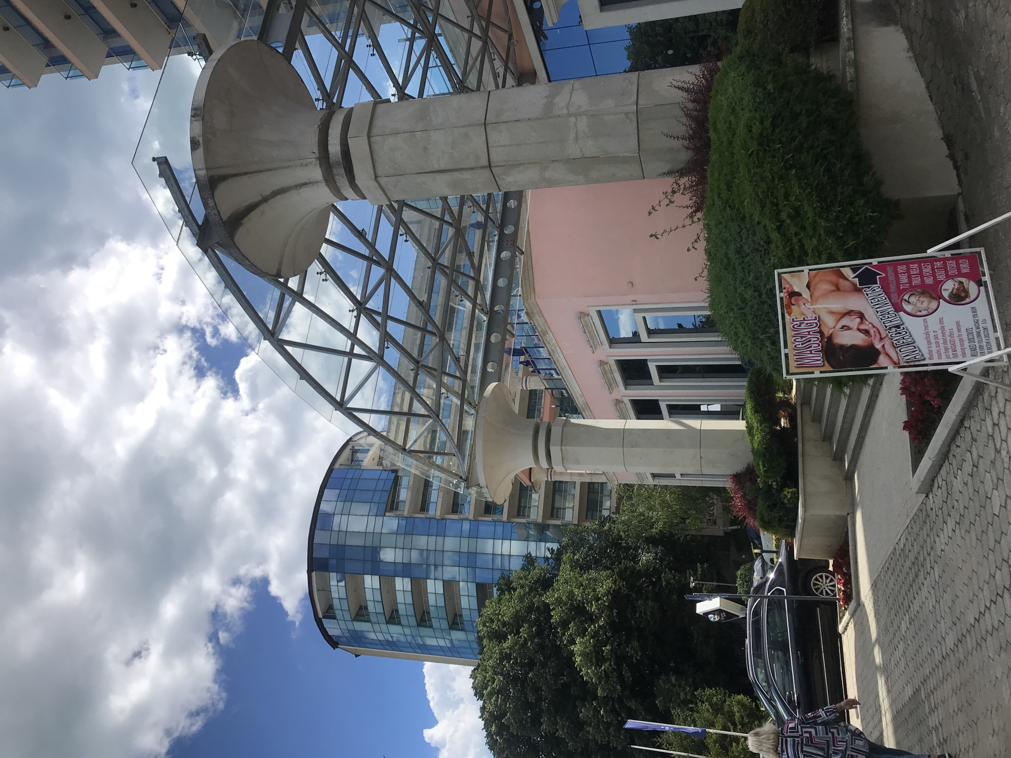 A scene unfolds in front of a modern building complex, likely a hotel or resort, located in Varna, Bulgaria. The architecture is contemporary, with a tall, curved building featuring a facade of blue glass windows dominating the background. In the foreground, a covered walkway with a distinctive glass roof supported by large, sculpted concrete pillars leads to a pastel pink building.
Several elements suggest a tourist-oriented environment. A sign advertising "MASSAGE AND FACE TREATMENTS" is prominently displayed on a stand. The sign features an image of a person receiving a massage and lists service details. Lush green bushes and flowering plants provide landscaping around the buildings, with red flowers visible in planters.
The ground is paved with interlocking bricks. A black sedan is parked on the side of the road, with two people walking away from the camera on the paved area. The sky above is a mixture of blue and white clouds, indicating fair weather. The overall impression is one of a well-maintained public space, possibly a hotel entrance or a recreational area in a coastal city.