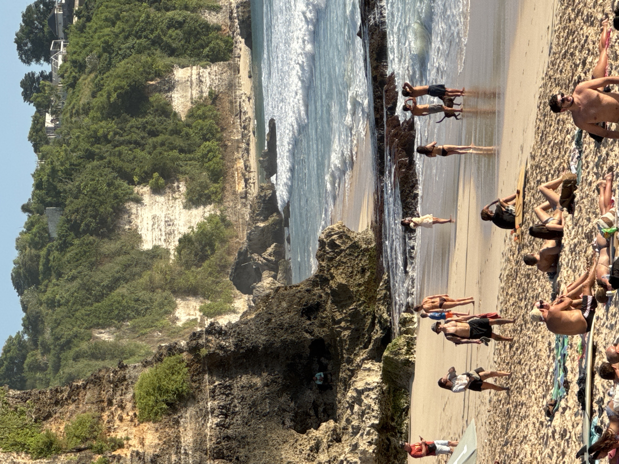 The image shows a beach scene in Kuta Selatan, Indonesia. Turquoise ocean waves crash against a rocky shore adjacent to a sandy beach where people are sunbathing, walking, and standing. Some people are closer to the water's edge. On the left, a cliff face is covered in lush green vegetation. In the background, more trees and buildings are visible.