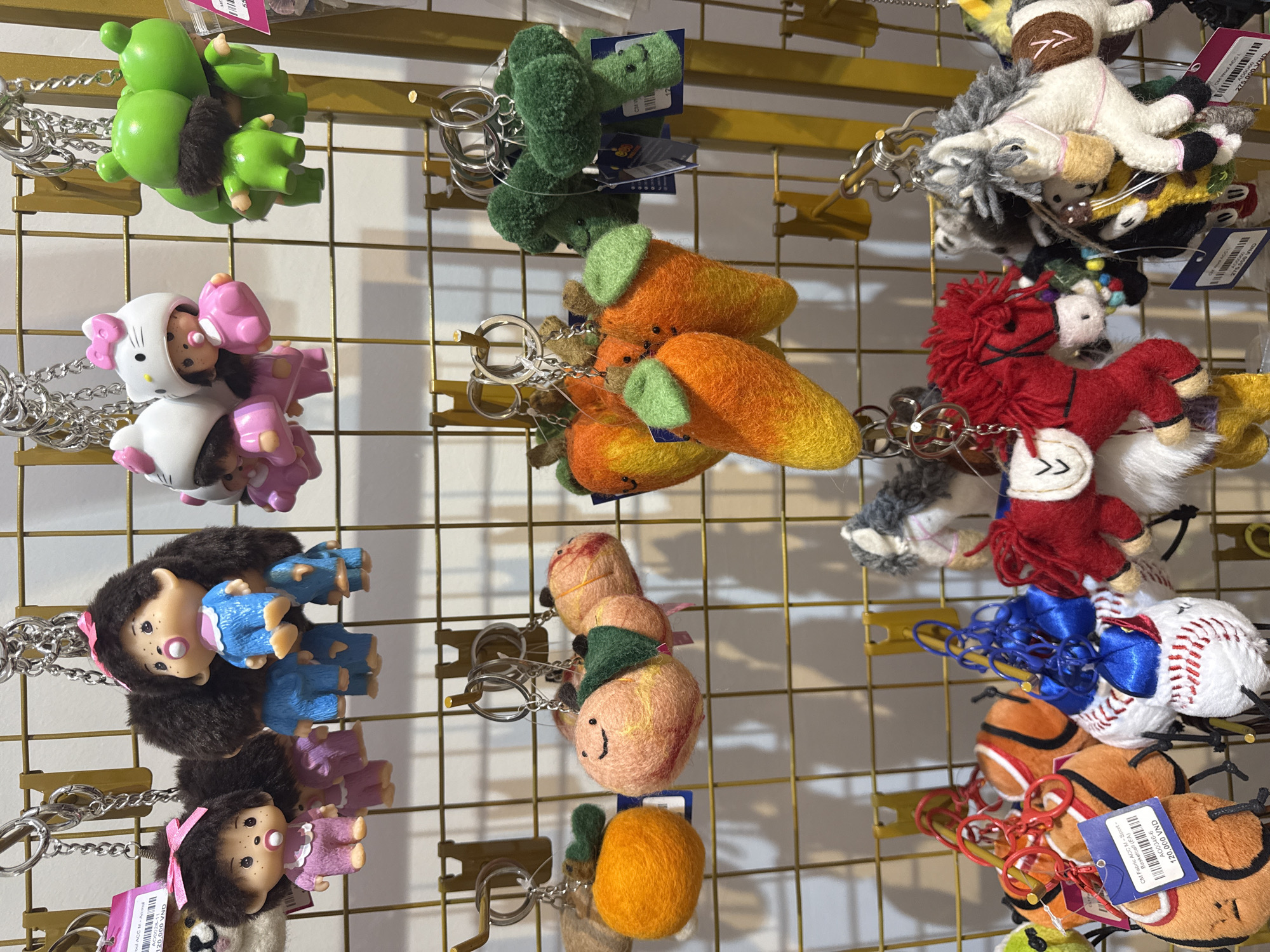 This is a close-up, eye-level shot of a display of various novelty keychains hanging on a gold wire grid. The keychains are diverse in design, featuring an assortment of plush and plastic characters.
In the top left, a cluster of bright green, frog-like figures with black fuzzy details. Below them is a white Hello Kitty-themed keychain with pink accents and multiple attached figures. To the right of the green frogs, a series of fuzzy green broccoli-like keychains are displayed. Below these are several orange carrot-shaped keychains. Further down the grid, a group of needle-felted peaches with smiling faces are visible.
The right side of the display showcases a variety of plush horses in different colors, including white with grey manes, and a red horse. There are also keychains resembling baseballs and what appear to be basketballs or tennis balls. A few keychains have small colorful bows attached.
The setting appears to be a retail environment, possibly a souvenir shop or market stall, given the grid display and the presence of price tags. One visible price tag reads "120,000 VND" and indicates "Basketball (EA)". Another tag reads "275,000 VND". The lighting is bright and even, suggesting an indoor setting during daylight hours. No people are visible in the frame. The overall impression is a colorful and whimsical collection of small decorative items.