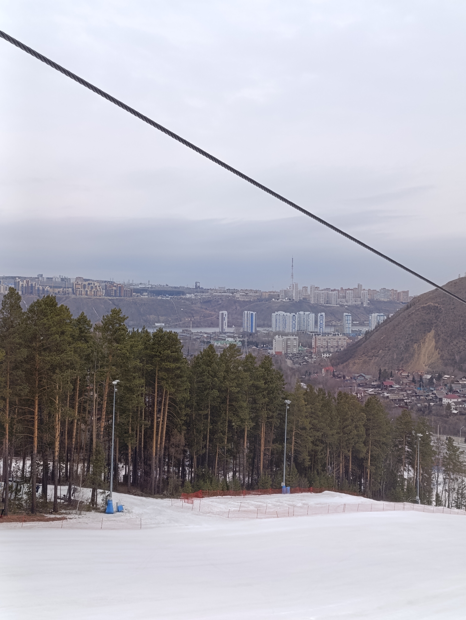 The image shows a ski slope in Krasnoyarsk, Russia. The foreground is dominated by a snowy slope, with a line of trees bordering the top of the slope. Orange safety netting is visible along the edge of the slope. Streetlights are placed among the trees. A cable is visible across the top of the image, suggestive of a ski lift. In the background, a city skyline is visible under an overcast sky. The architecture of the buildings in the skyline suggests that this is likely a modern city. The scene suggests a cold weather, winter recreation setting within an urban environment.