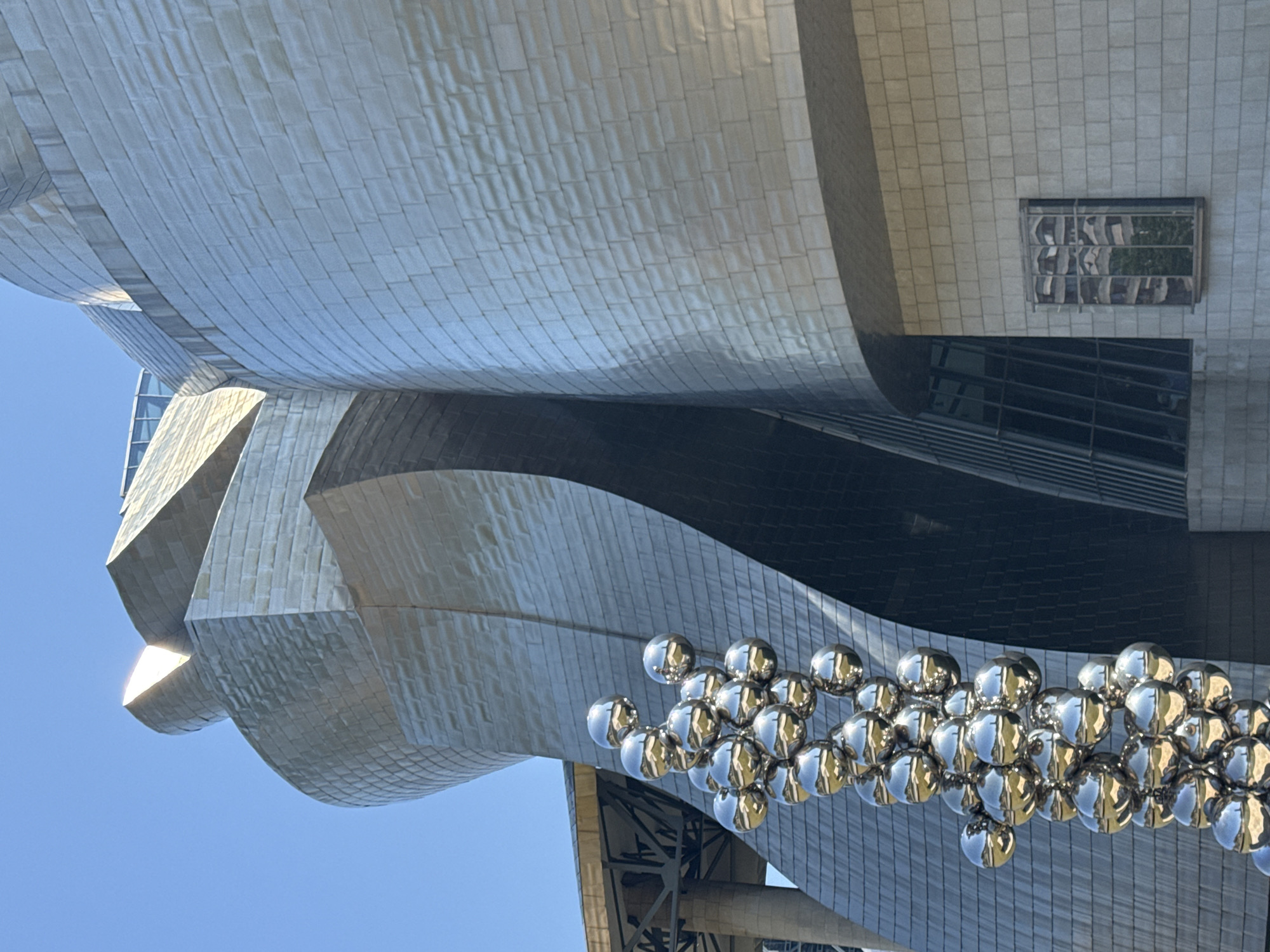 A low-angle, outdoor, close-up shot captures the exterior of the Guggenheim Museum Bilbao, Spain. The iconic titanium-clad building features curved, wave-like structures. In the foreground, a large, reflective sculpture composed of numerous metallic spheres is prominently displayed. The sky above is clear and blue.