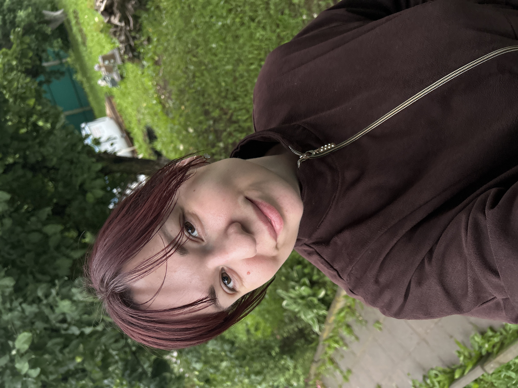 This media file contains a selfie of a person with reddish-brown hair. The person is wearing a dark brown hooded jacket with a silver zipper. Their face is turned towards the camera, and they appear to be smiling slightly. The background is blurred, showing greenery, including trees and grass. There are also some hints of a paved pathway and possibly some structures in the distance. The lighting suggests it is daytime, and the greenery implies an outdoor setting, likely a garden or park. The overall mood seems relaxed and casual. No text is visible in the image. The location context provided is Фоки, Russia.