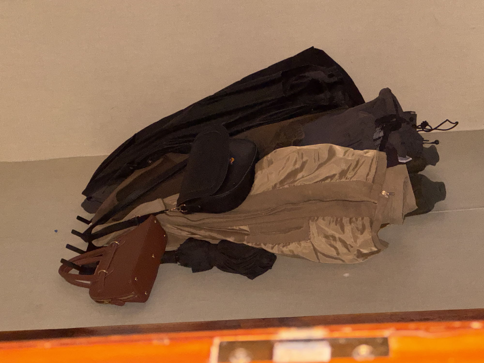 In the city of Yarominski Sel'ski Sovet, Belarus, a collection of outerwear and accessories hangs on a wall-mounted rack. A brown, structured handbag with twin handles is positioned on the leftmost hook. To its right, a black umbrella is folded and suspended. A light brown, quilted jacket is draped over the rack, its zipper partially visible. A black crossbody bag with a flap closure hangs next to the jacket. Further to the right, several dark-colored jackets or coats are present, their forms obscured by their arrangement on the hooks. The background consists of a neutral-colored wall. The scene is captured with artificial lighting, casting soft shadows. The orange-toned frame of what appears to be a door or window is partially visible on the left edge of the image.