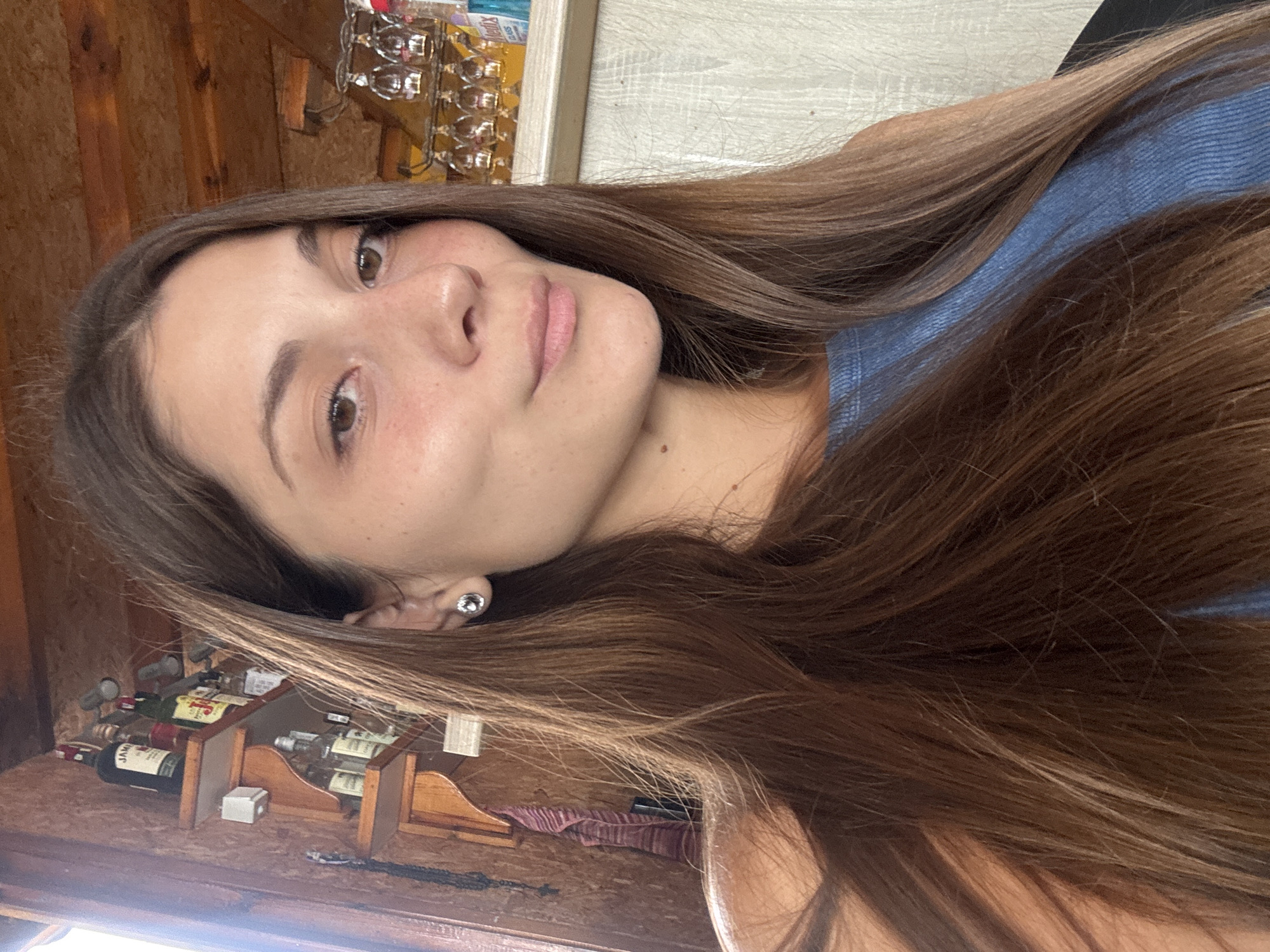 A young woman with long, brown hair is looking at the camera. She is wearing a blue, ribbed top. In the background, there is a wooden shelf with several bottles of alcohol, and a rack of wine glasses. The overall setting appears to be a bar or restaurant in Sozopol, Bulgaria, given the rustic wooden interior and the selection of alcoholic beverages. The lighting suggests it is daytime, possibly indoors near a window due to the bright light on the left side of the image.