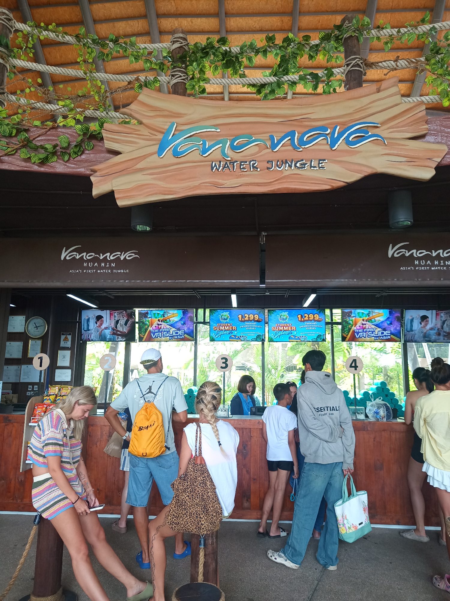 Here is a detailed description of the image:
The image shows a scene at the entrance to Vananava Water Jungle in Hua Hin City Municipality, Thailand. There are people queuing at ticket counters numbered 1 to 4. The ticket booths are under a covered area. A sign above the booths displays “Vananava, Hua Hin, Asia's First Water Jungle.” Above the sign, a wooden structure with "Vananava WATER JUNGLE" is seen, decorated with foliage and rope.
Several individuals are present. At booth 1, a woman with blonde hair is looking at her phone, seated on a wooden post. Next to her is a man with a light-colored hat and an orange backpack who is facing the ticket booth. Beside him, a woman with braided hair is facing the booth with a leopard print bag. At booth 3, a staff member is attending to customers. A boy in a white t-shirt is facing the ticket booth and next to him stands a person wearing a gray sweatshirt with “ESSENTIALS” written on it. At booth 4, a group of people are waiting in line.
Above the ticket booths are TV screens displaying advertisements and ticket information, including prices of "Student Summer Splash" tickets at 1,299.00.
The environment is bright, with natural light illuminating the scene. There are wooden structural elements indicating the entrance to a recreational facility. The overall setting is casual and indicative of a tourist attraction.