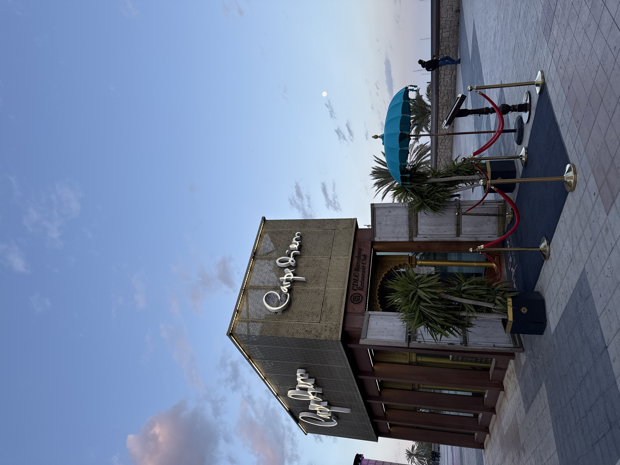 The image shows the exterior of the Carpe Diem Lounge Club (CDLC) in Barcelona, Spain. The building has a modern design with dark wood and metal accents. "Carpe Diem" is prominently displayed in illuminated script on the exterior. A blue parasol and stanchions with red velvet ropes mark the entrance. A person is walking in the distance on the promenade, which is paved with stone tiles. The sky is a pale blue with some clouds. A bright moon is visible.