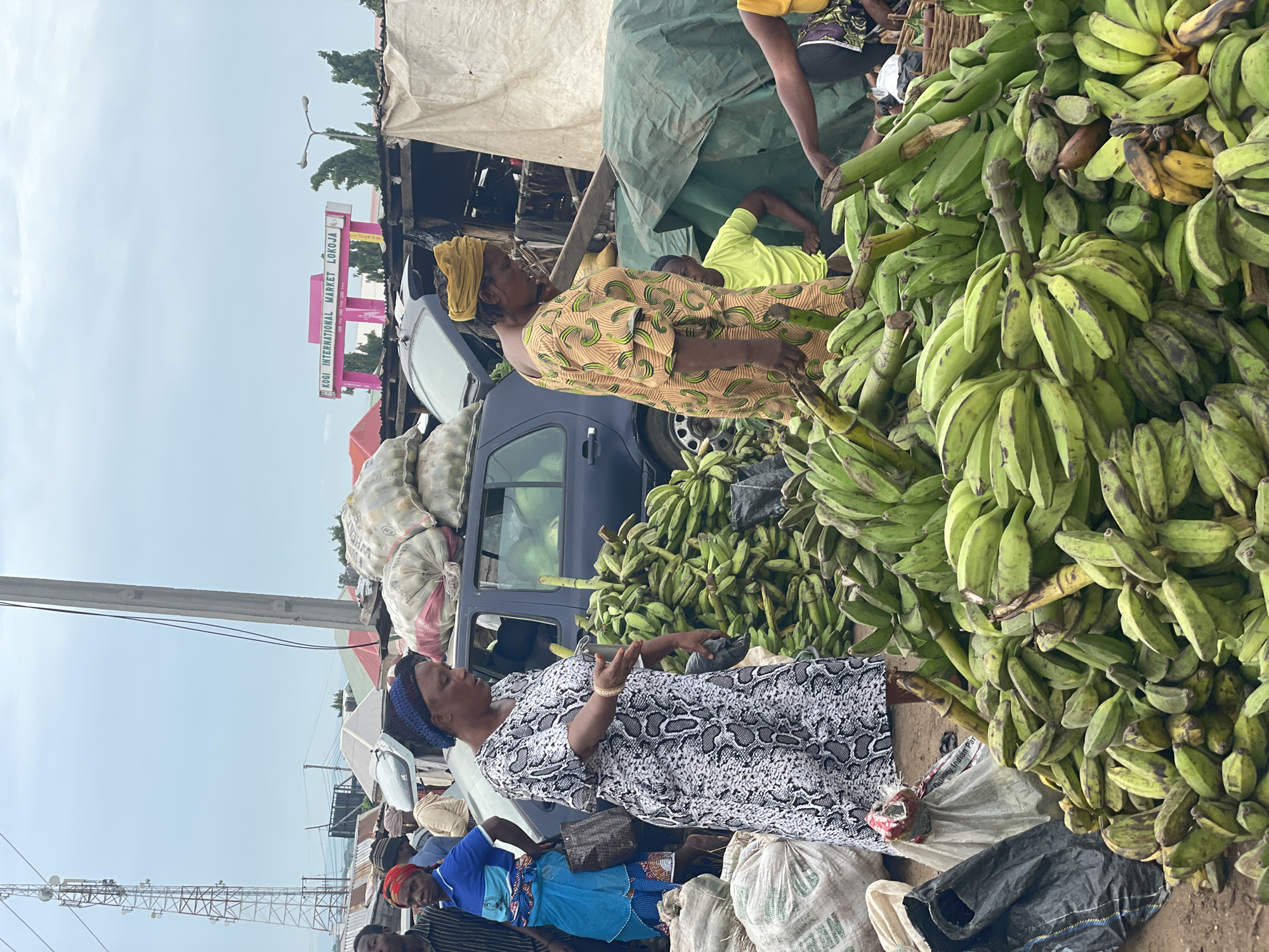 In Lokoja, Nigeria, a busy marketplace is depicted, with a focus on a large display of green bananas. Two women are prominently featured. One woman, wearing a black and white patterned dress and a blue headwrap, is standing in front of a dark-colored car and gesturing with her right hand, holding a black plastic bag. The other woman, in a yellow headwrap and a patterned orange dress, stands to the right of the bananas, looking down. Behind them, several large sacks, some filled with produce, are visible, along with a dark blue car with its trunk open. A bright pink sign reading "KOGI INTERNATIONAL MARKET LOKOJA" stands in the background, confirming the location. The scene is outdoors, under an overcast sky, with other people and market stalls also present.