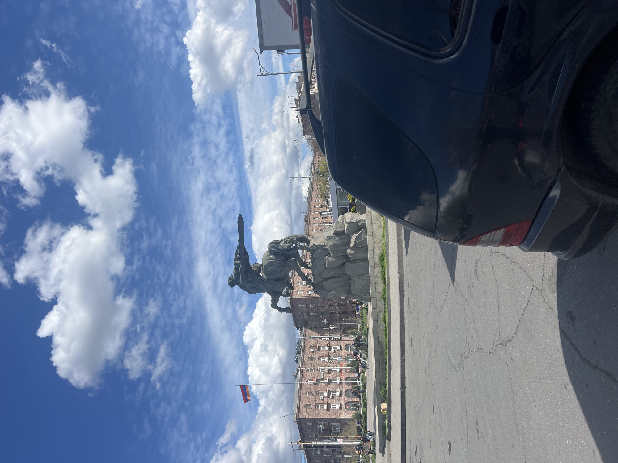 The image shows the statue of David of Sasun in Yerevan, Armenia, partly obscured by the rear of a black car on the right. The statue depicts a mounted warrior, sword raised, on a rocky pedestal. Behind the statue is a multi-story building with numerous windows and a flag of Armenia waving in front of it. The sky is blue with scattered white clouds. The scene is captured from street level, and a section of the asphalt roadway is visible in the foreground.