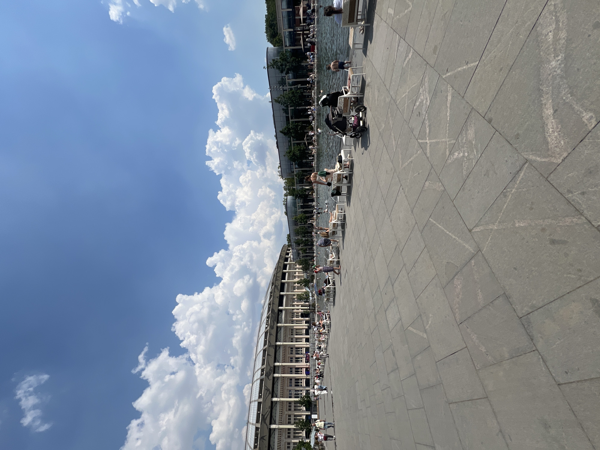The image depicts a wide paved area in front of a large, modern stadium in Moscow, Russia. The stadium, with its distinctive roof structure and white facade, dominates the left side of the frame. A body of water, possibly a fountain or reflecting pool, is situated in the middle ground, with people relaxing on lounge chairs around its perimeter. Some individuals are standing or walking on the paved area, while others are in or near the water. A baby stroller is visible on the right side of the frame. The sky is blue with scattered white clouds. The paving stones are large and grey, with some faint markings or scratches visible. The overall scene suggests a public space during a warm, sunny day.