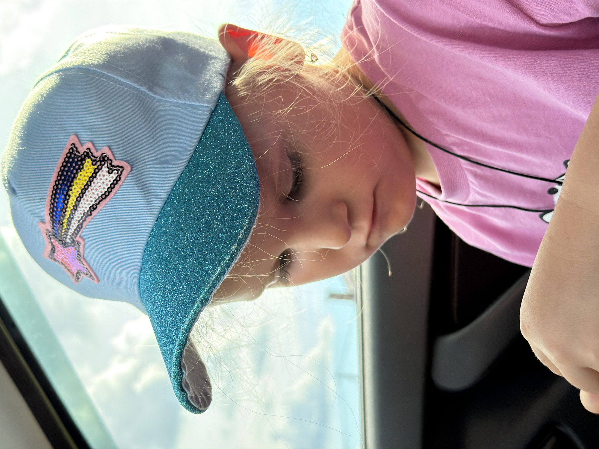This is a close-up, low-angle, upside-down outdoor photograph of a young child wearing a light blue baseball cap with a sparkly turquoise brim and a colorful embroidered patch of what appears to be a lightning bolt or a stylized rainbow. The child has fair hair, and their face is turned downwards, partially obscured by the cap and their hair. They are wearing a bright pink t-shirt. The background consists of a bright sky with some clouds, suggesting it is daytime. The lighting is strong, casting some highlights on the child's hair and the cap. The child's hand is visible, positioned near the pink t-shirt. The scene appears to be an outdoor setting, possibly in a vehicle given the dark interior elements visible at the bottom right of the frame. There is no visible text in the image. The photograph was taken in Chaikovsky, Russia.