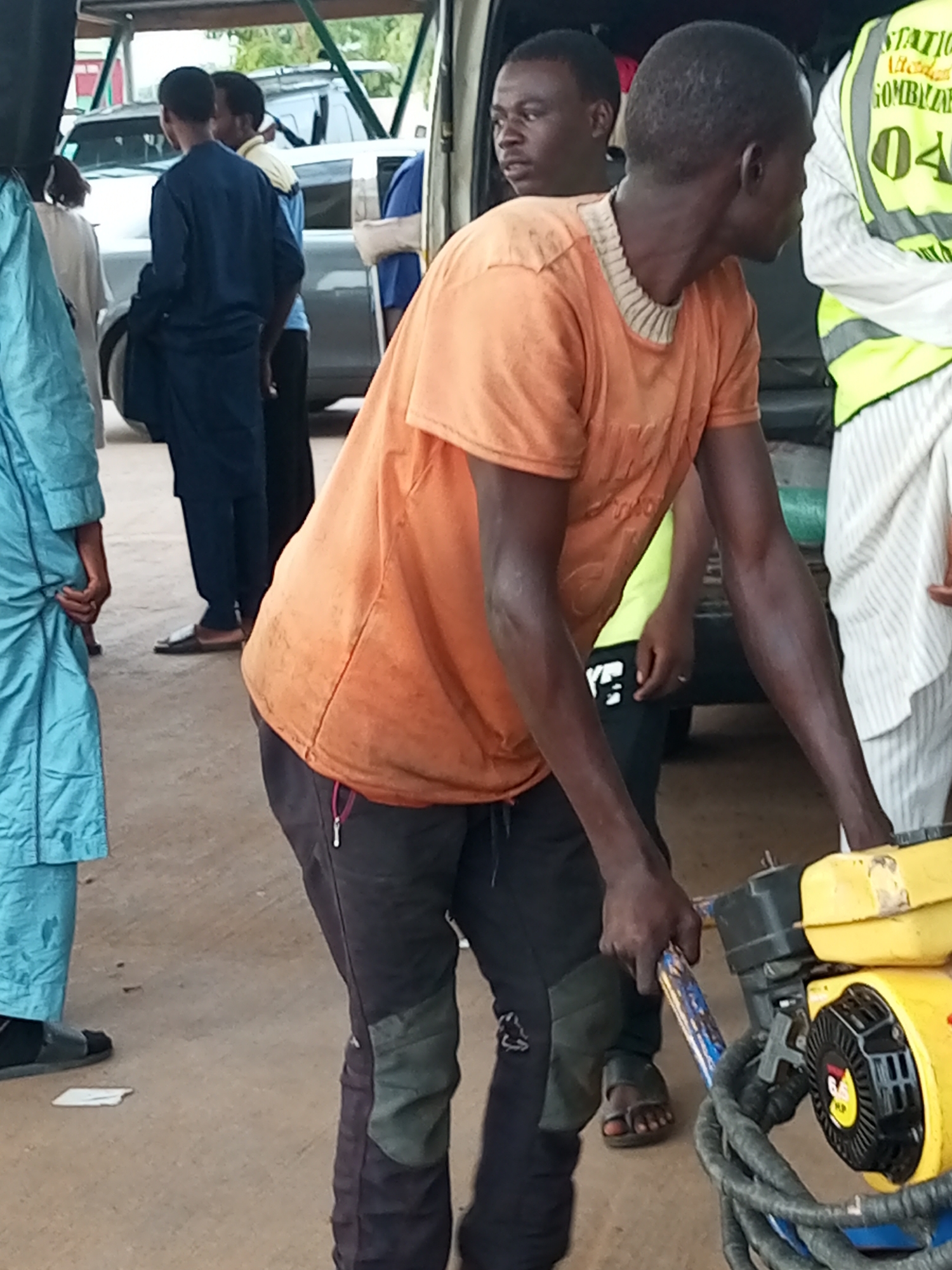 The image depicts an outdoor scene in Gombe, Nigeria, possibly near a road or a market area. In the foreground, a man wearing an orange t-shirt and dark pants is operating a yellow and black piece of equipment, which appears to be some sort of compaction tool or generator. His hands are on the handle of the equipment. Behind him, another man wearing a yellow safety vest with the inscription "STATION 040" is visible. To the left, a person in a light blue garment is also present. In the background, a white vehicle, possibly an SUV, is parked. The ground is paved with concrete or asphalt, and there are hints of foliage or trees in the distance. The overall lighting suggests it is daytime. The people are of African descent.