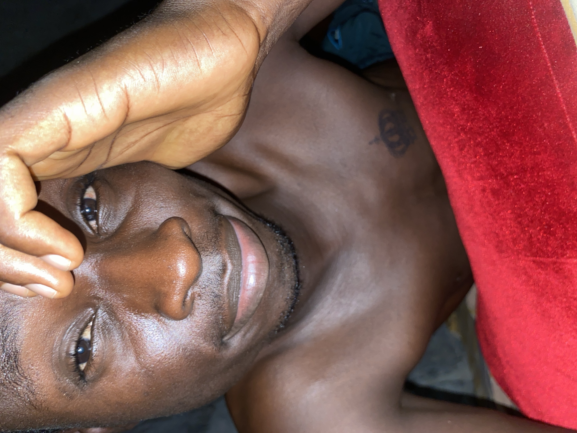 The image shows a close-up, angled view of a shirtless Black man lying down. He is looking up towards the camera, with his right hand partially covering his forehead. A small tattoo of a crown is visible on his chest, near his shoulder. A red, textured object, possibly a blanket or pillow, is to his right. The lighting is subdued. There is no explicit information to indicate the location is Narayi, Nigeria.