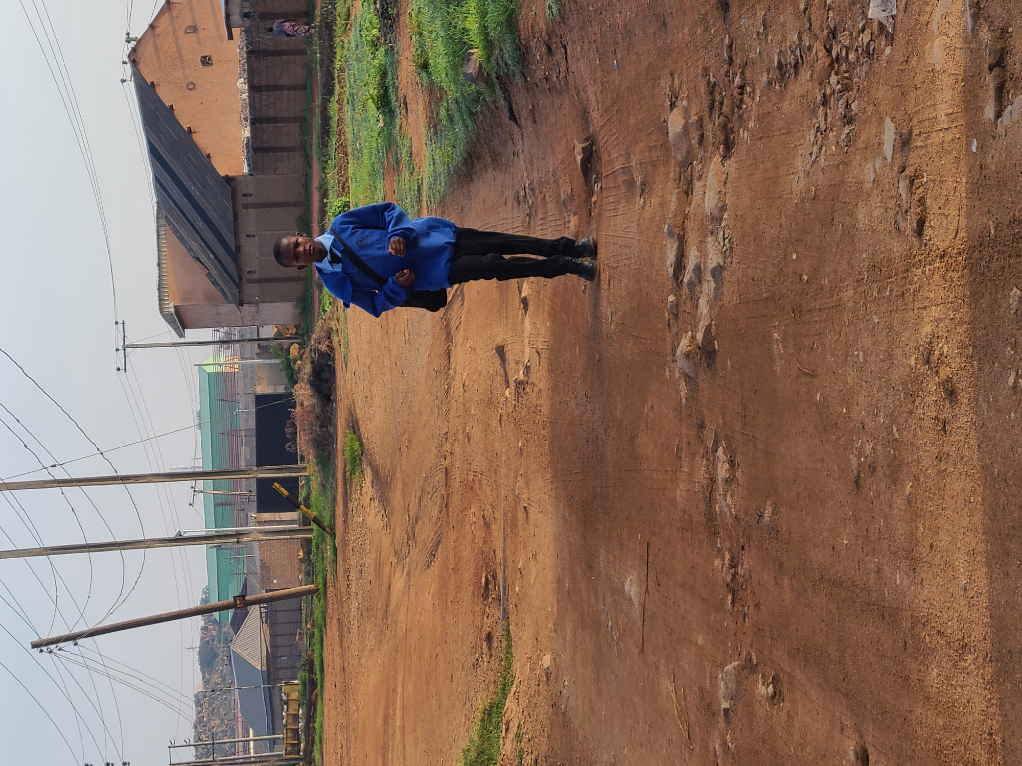 A young boy walks on a dirt road in Jos, Nigeria. He is wearing a blue sweater, black pants, and a black bag over his shoulder. The road is unpaved and has scattered rocks. In the background, there are power lines, buildings, and some vegetation. The sky is clear and the lighting suggests it is daytime.