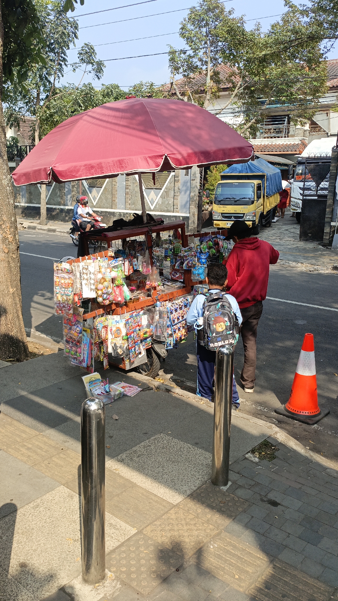 The image captures a street scene in Salatiga, Indonesia, on a sunny day. A mobile cart, laden with a variety of colorful toys and small items, is parked on the sidewalk. The cart is covered by a large, vibrant red umbrella. Two boys are standing near the cart, one of whom is wearing a backpack with a cartoon character design. A man in a red hoodie stands behind the boy with the backpack. In the background, a yellow truck with a blue tarp is parked on the street, and other vehicles are visible further down the road. Houses and trees line the street, suggesting a residential or mixed-use area. The sidewalk features textured paving tiles, typical for pedestrian walkways. An orange traffic cone is placed on the side of the road, possibly indicating a temporary hazard or traffic diversion. A person on a scooter is seen in motion on the street, adding to the dynamic nature of the scene.