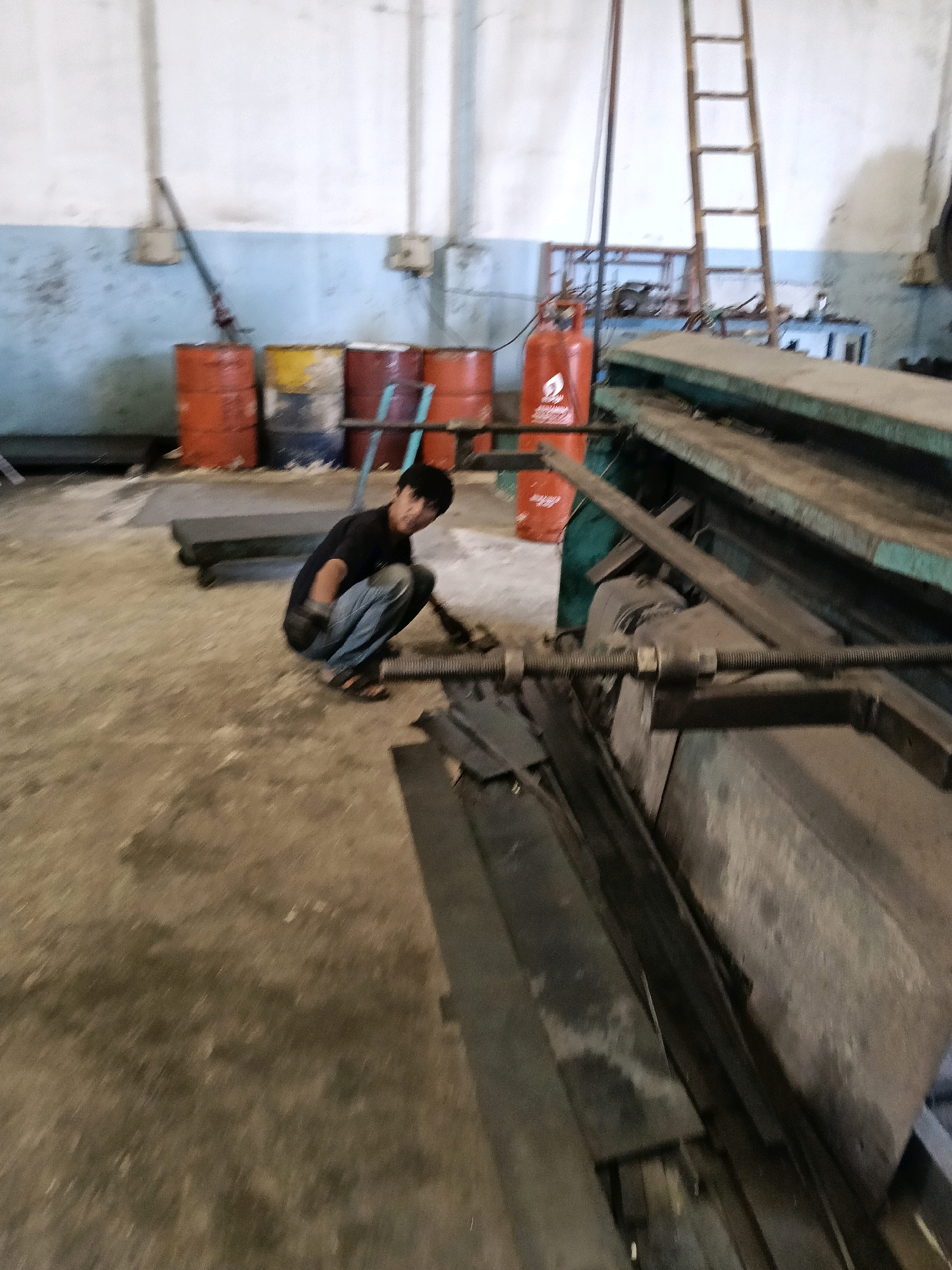 In a dimly lit industrial workshop, a person is crouched on the dusty concrete floor, wearing a dark t-shirt and blue jeans. The individual is facing towards a large, green metal machine that dominates the right side of the frame. The machine appears to be a piece of industrial equipment, possibly a shear or press, with various metal components and a threaded rod visible.
In the background, several large, rusty barrels are stacked, with colors including orange and yellow. A red gas cylinder with a white logo is also present. To the far right, a wooden ladder is propped against a white wall, which has a blue stripe running across it. The overall environment suggests a working area with tools and materials typical of a metal fabrication or repair shop. The image is slightly blurred, indicating movement during capture. The location is identified as Belimbing, Indonesia, based on the context provided.