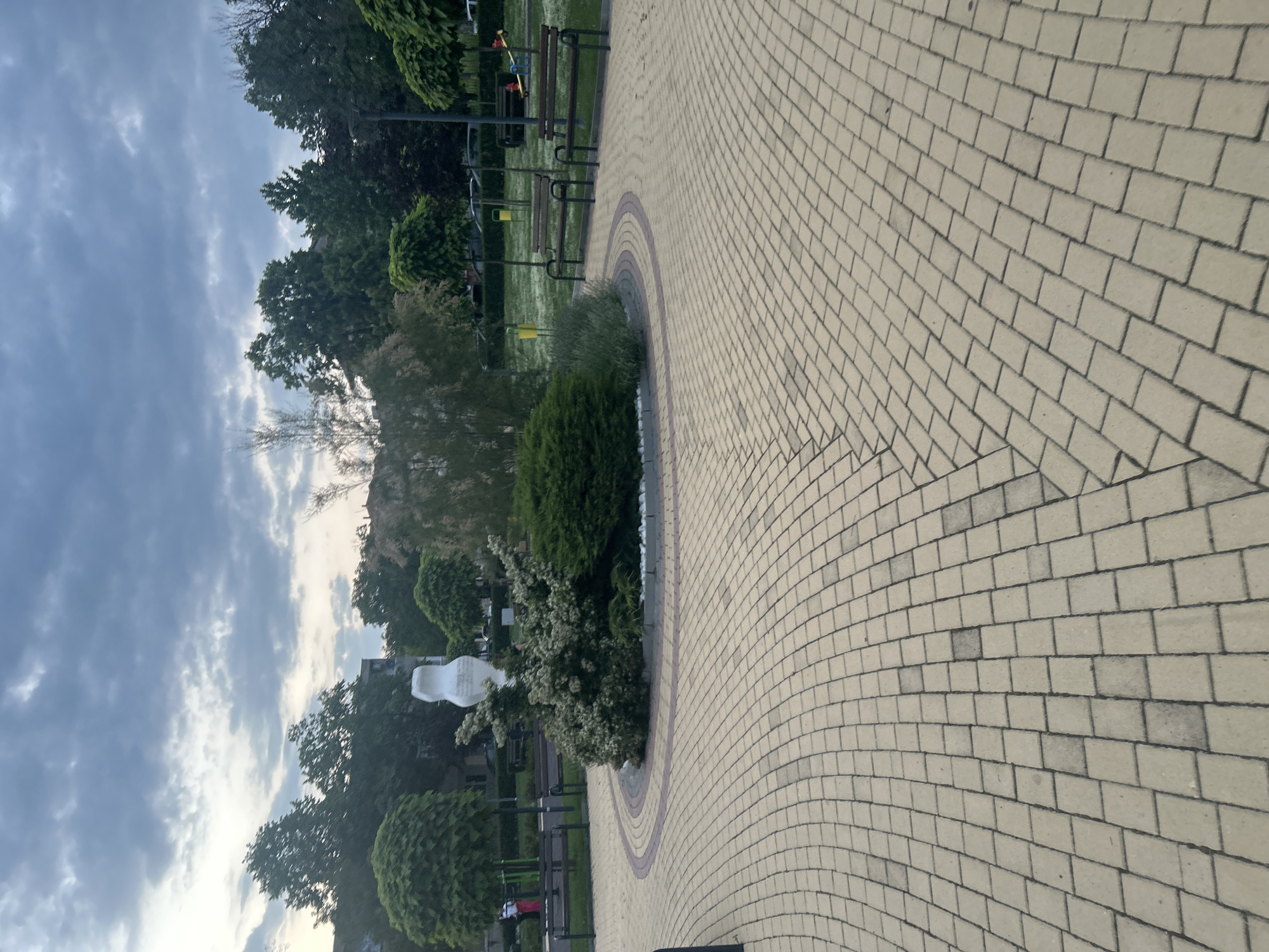 The image captures a peaceful public park or town square in Dzhebel, Bulgaria, under an overcast sky. A significant portion of the foreground and midground is dominated by a meticulously paved pathway made of light beige rectangular pavers. This pathway features an intricate pattern, including a large semi-circular section bordered by darker, reddish-brown pavers, which outlines a landscaped green island.
Within this landscaped island, various shrubs and bushes are visible, some neatly manicured, others with white blossoms. A prominent, abstract white sculpture, with smooth, organic curves, stands among the greenery, serving as a focal point.
Beyond the paved area and the central island, a well-maintained green lawn stretches out, featuring several dark wooden park benches with metal supports, inviting visitors to rest. Further back, numerous trees with dense green foliage, including both deciduous and coniferous types, form a lush backdrop, partially obscuring the distant horizon and any potential buildings. The cloudy sky, with its soft, diffused light, suggests either early morning or late afternoon. The scene is devoid of people, contributing to an overall tranquil and serene atmosphere. No visible text is present in the image.