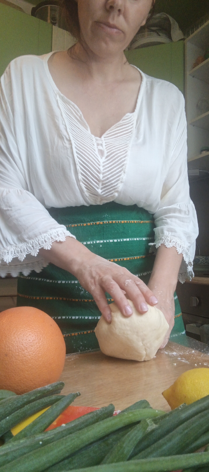 A woman is standing in a kitchen, wearing a white peasant-style blouse and a green apron. Her hands are covered in flour as she kneads a ball of dough on a wooden countertop. In front of her, arranged on the counter, are a grapefruit, a lemon, and several stalks of green onions, with hints of yellow and red peppers also visible. The background shows kitchen cabinets and shelves, suggesting a domestic setting.