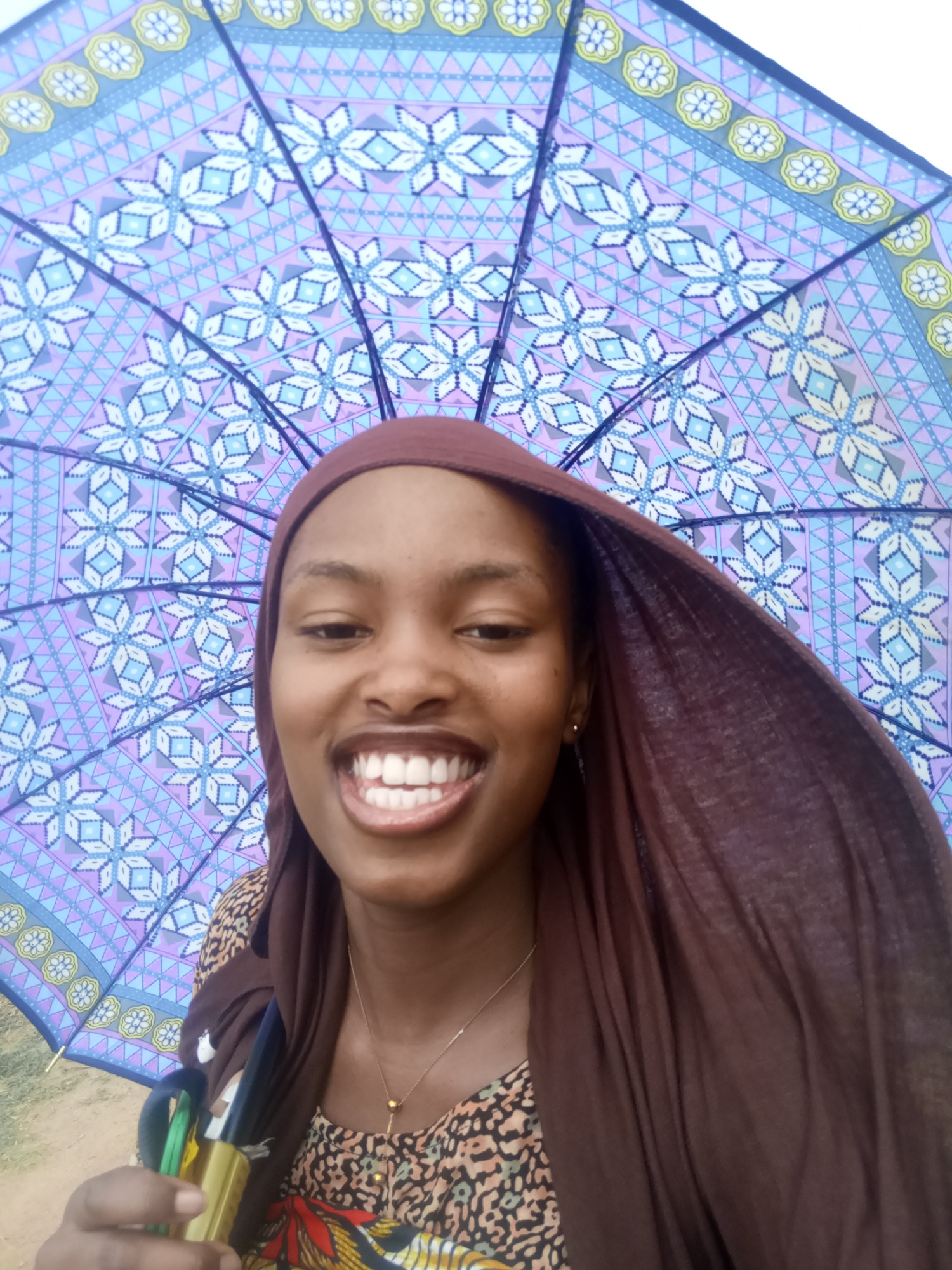 A smiling woman with dark skin and a brown head covering is holding a closed umbrella with a blue, lavender, and white geometric pattern. She is wearing a patterned shirt with reddish-orange, yellow, and black elements and a gold necklace. The location appears to be outdoors, possibly in Unknown, Unknown.