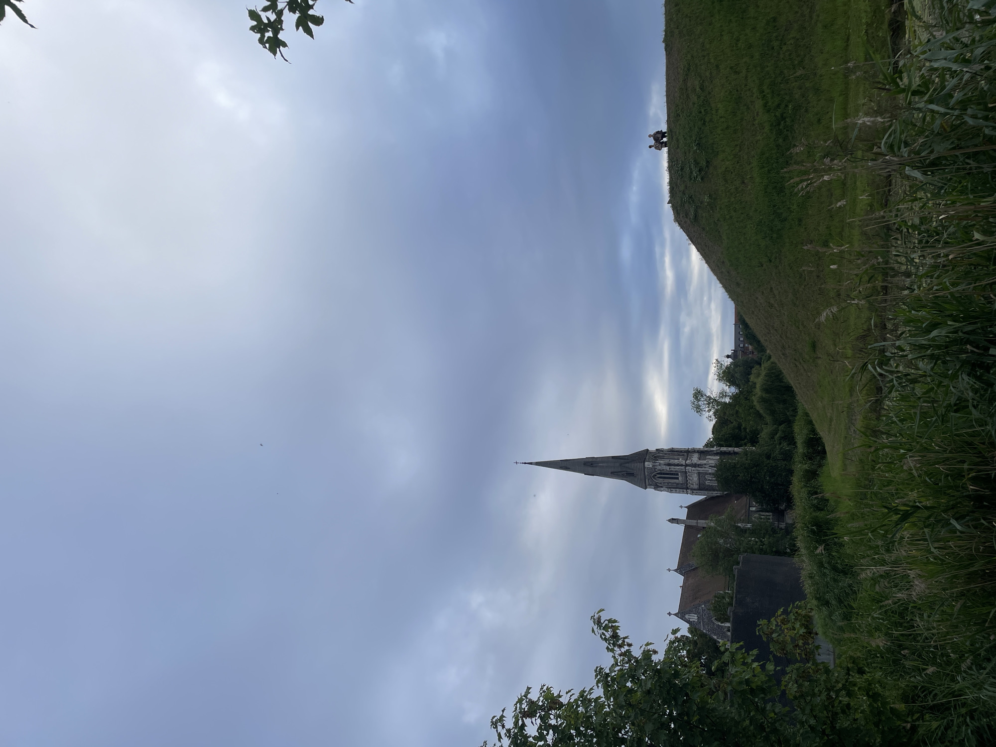 The image depicts St. Alban's Church in Copenhagen, Denmark, seen from a grassy knoll. The church, with its Gothic Revival architecture and tall spire, is situated amidst lush greenery. Two individuals are visible on the crest of the hill, appearing to be conversing. The sky is overcast with dramatic clouds, suggesting either early morning or late afternoon. The foreground is dominated by tall grasses and reeds, hinting at a body of water nearby. The overall scene is tranquil and picturesque, combining architectural grandeur with natural beauty.