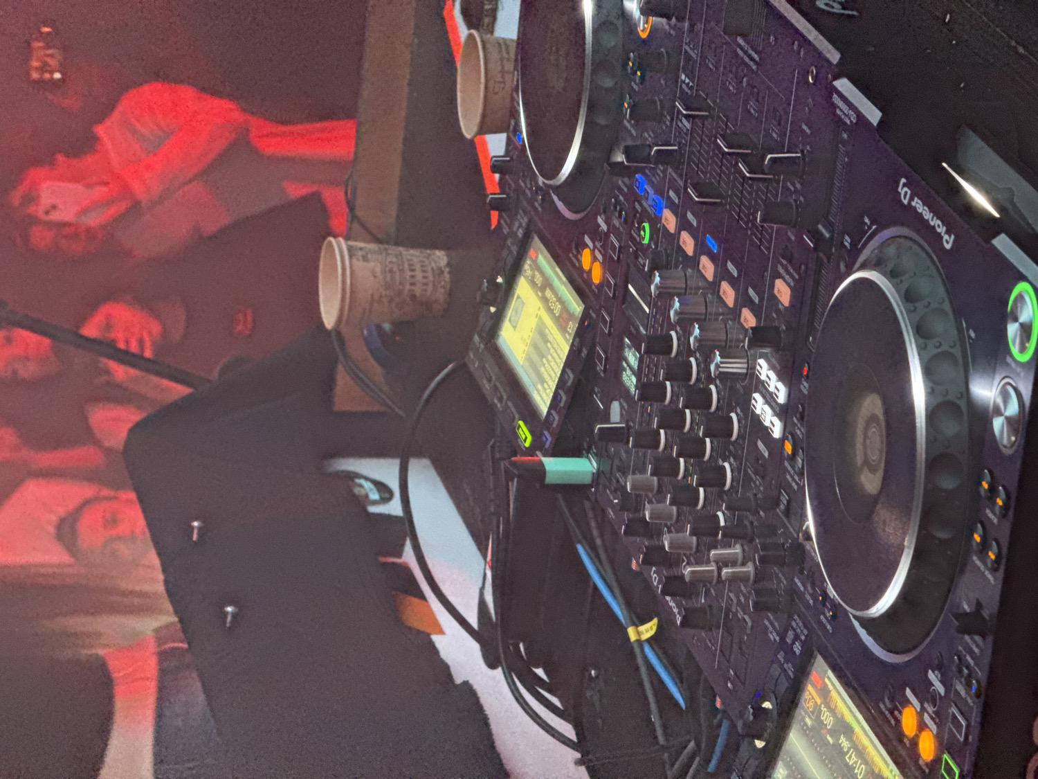 The image captures a vibrant, low-light scene from a club or bar in Vladivostok, Russia, showcasing a professional DJ setup and an energized crowd. The primary subject is a dark-colored Pioneer DJ controller and mixer, positioned at an angle, displaying numerous illuminated buttons, faders, knobs, and two jog wheels. Two small digital screens on the equipment show timecodes like "00:55:00" and "01:47:00," along with other track information that is not fully legible.
To the left of the DJ equipment, two disposable cups, possibly paper, are placed on a dark surface, one featuring a faint, repeating pattern. Various cables are visible, connecting components of the setup. The background reveals a crowd of people, illuminated by a dominant red artificial light, creating a hazy and atmospheric glow. Their faces and figures are largely obscured due to the lighting and blur, but they appear to be engaged in the event, with one person in the upper left seemingly holding a phone.
The scene suggests a nighttime event, likely a party or musical performance where a DJ is actively playing music. The overall mood conveyed is one of entertainment and nightlife. No specific weather details are discernible, and location cues beyond the club setting are not visible in the image itself, though the context given is Vladivostok, Russia. The text "Pioneer DJ" is clearly visible on the equipment.