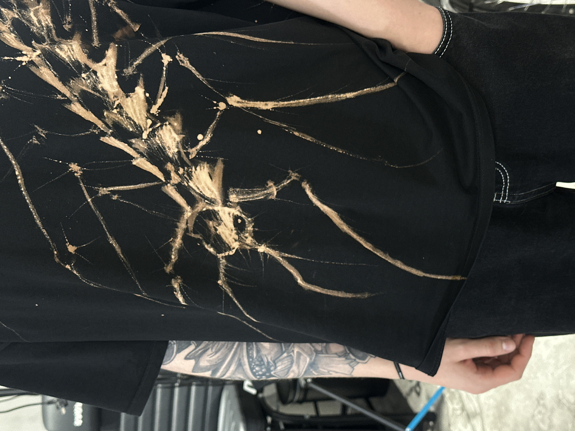 The image shows a person wearing a black t-shirt with a bleach-splattered design resembling a spider. The person is also wearing dark jeans. A portion of an arm with a tattoo is visible. In the background, part of a chair and other indiscernible objects are visible. There is no indication in the image that the location is Moscow, Russia.