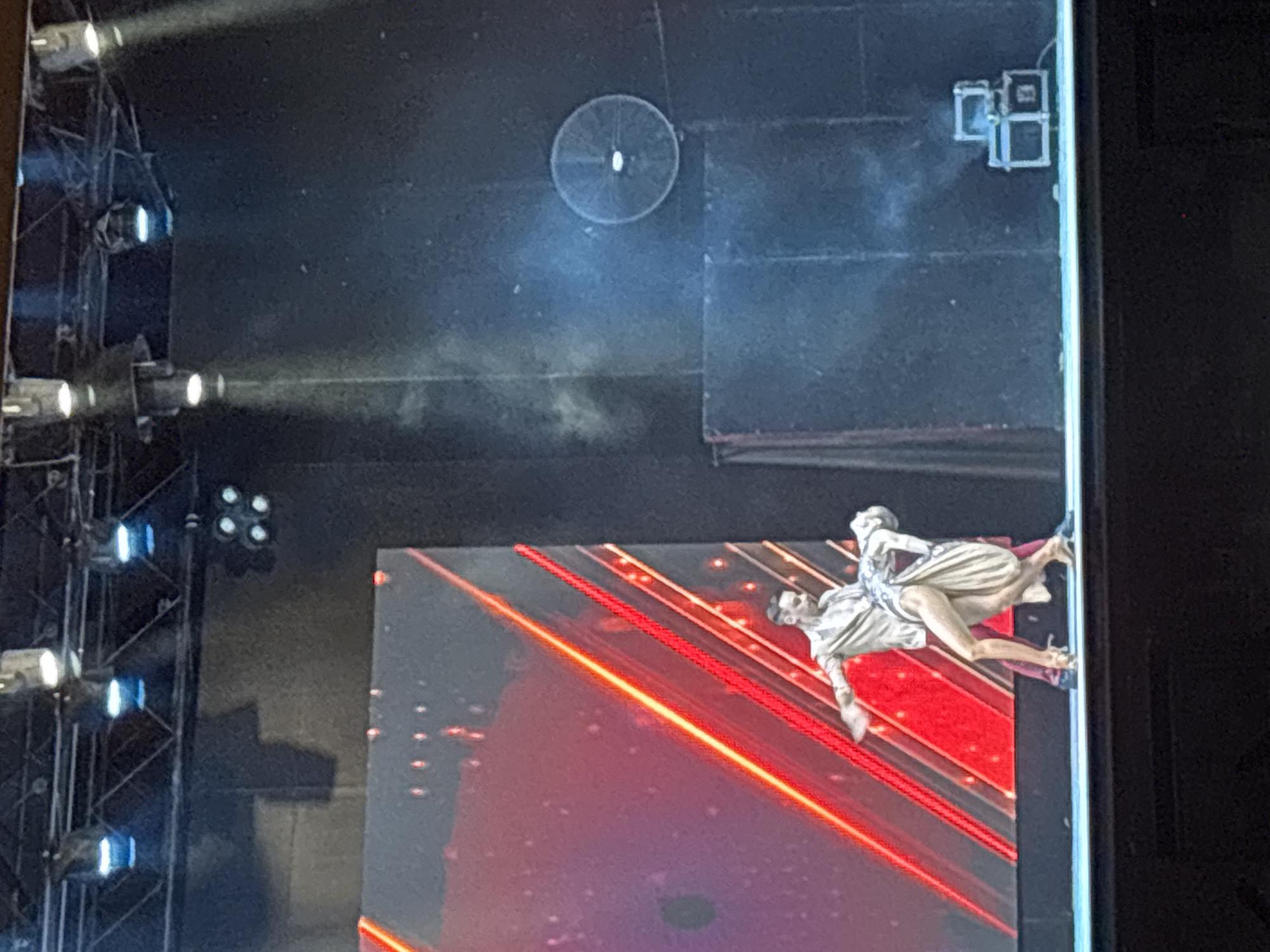 Two dancers, a man and a woman, are performing a ballroom dance routine on a stage in front of a large LED screen displaying red and yellow diagonal lines. The man is wearing a light-colored shirt and dark pants, while the woman is in a tan dress. The stage is lit by spotlights from above, and there is some haze or smoke in the air. In the background, a circular fan is mounted on the dark backdrop, and two stacked equipment cases are visible. The scene appears to be an indoor event or performance.