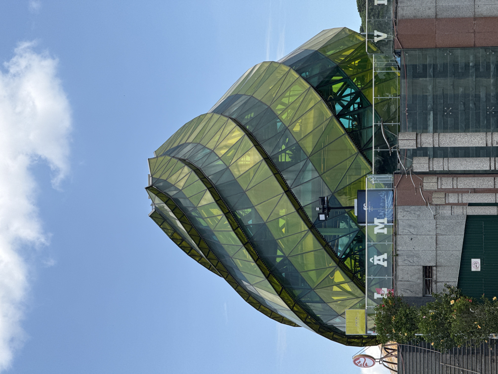 The image captures a unique architectural structure, the The Cinema Centre in Da Lat, Vietnam. The building features a distinctive green, layered exterior resembling stacked, curving slices. The glass facade is patterned with triangular divisions, giving it a multifaceted look. The building is partially obscured by a vertical sign spelling out "LÂM ĐỒNG" in large white letters. Adjacent to the structure is a standard grey concrete building. Below the unique architecture, there's signage for "mobile city" and "galaxy cinema". Vegetation, including a bush with roses, is visible at the base of the building. A partially obscured sign is seen below the bush. The sky above is clear blue with scattered white clouds. The image is oriented vertically.