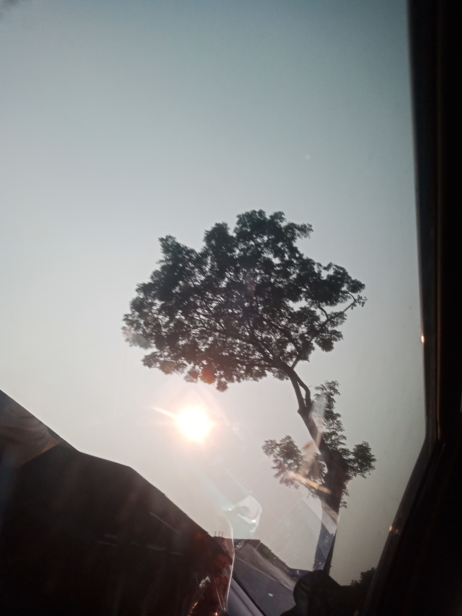 The image shows a view through a window, likely from a vehicle, with a tree silhouetted against a hazy sky. The sun is visible as a bright lens flare, casting a warm glow. Reflections on the window obscure parts of the scene, including what appear to be other vehicles or structures. The bottom left of the image shows a dark, angled surface that could be part of the vehicle's interior or an external object. The sky is a pale, washed-out blue, suggesting either early morning, late afternoon, or overcast conditions. The general atmosphere is soft and diffused. The specific location in Tangerang, Indonesia, cannot be determined from the visual information presented in the image.