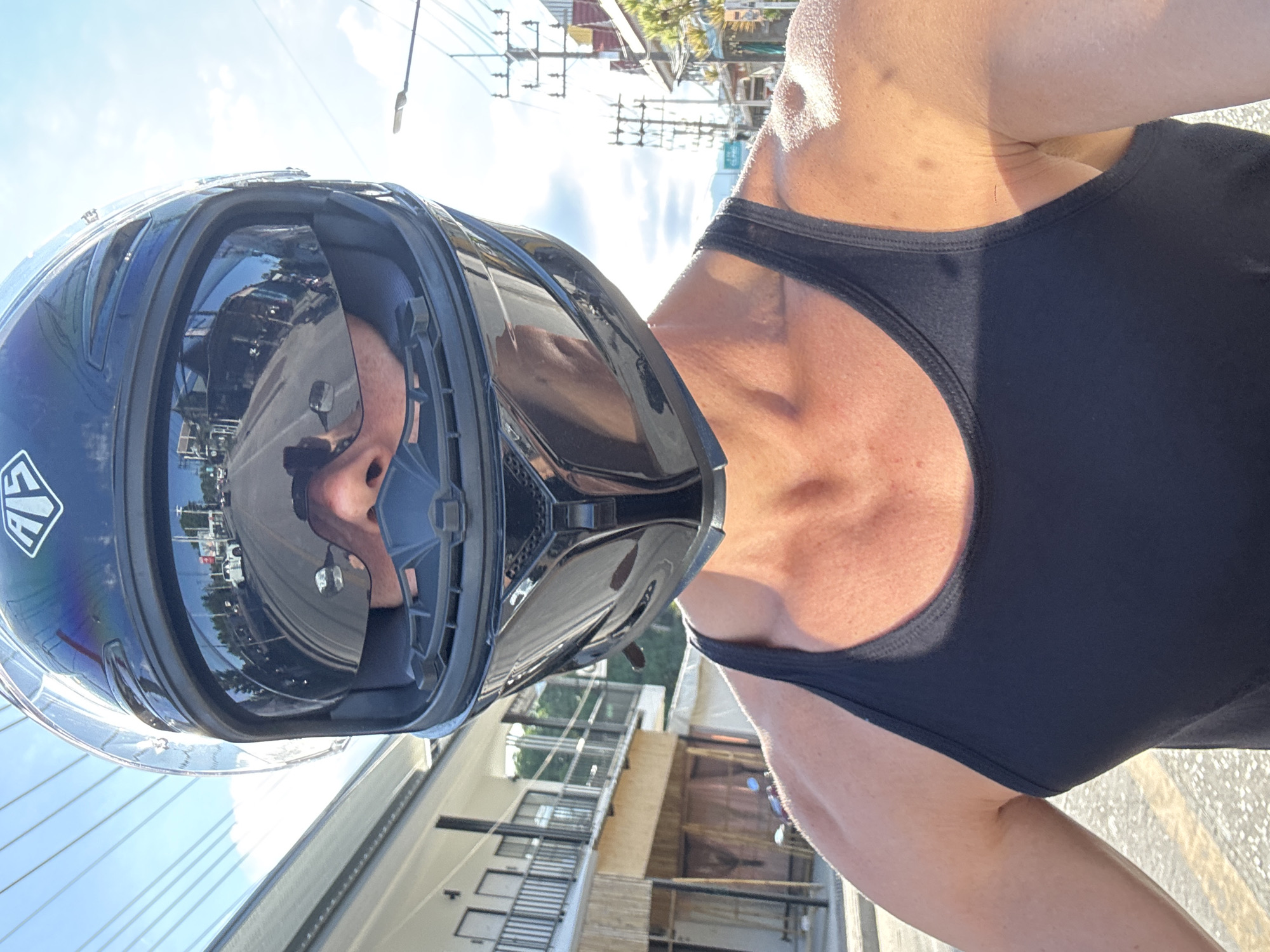 A close-up, first-person perspective shot shows a person wearing a black full-face motorcycle helmet with a reflective visor, obscuring most of their face. The visor reflects a street scene with buildings on both sides, utility poles, and vehicles, indicating a location in a populated area. The person is wearing a black tank top, revealing muscular shoulders and neck. The background suggests a bright, sunny day with a clear blue sky and some clouds. The surroundings appear to be a street in a tropical or subtropical region, possibly Ko Pha Ngan, Thailand, with architecture and vegetation consistent with such a location. The road surface is visible, and the overall impression is that the person is on a motorcycle, moving through a street.