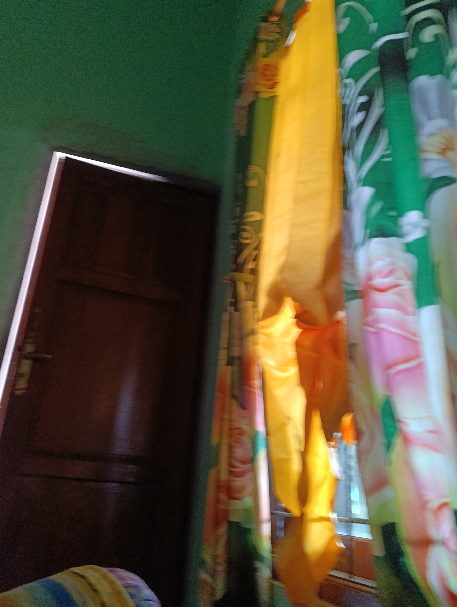 The image shows a dimly lit interior scene, likely a room. On the left, a dark brown door with a horizontal panel and a brass door handle is visible. The door is set against a green wall. To the right of the door, floral patterned curtains are draped, featuring green, yellow, pink, and white hues. The curtains appear to be partially drawn, allowing some light to filter through a window behind them. The overall impression is one of a domestic setting. There are no people visible in the image. The location is stated to be Jombor, Indonesia.