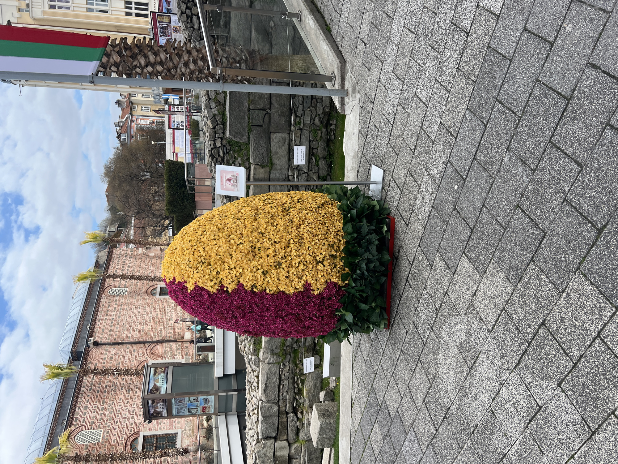 The image depicts an ornamental arrangement of flowers, predominantly yellow and purple, set against a backdrop of historical and modern architectural elements in Plovdiv, Bulgaria. The floral arrangement is egg-shaped, with yellow flowers comprising the upper portion and purple flowers the lower. It is bordered by green foliage.
Behind the flowers are visible stone ruins. Adjacent to the flowers and ruins is a modern building constructed of red brick with glass elements. A Bulgarian flag is visible at the upper left corner of the scene, suggesting the national context. A paved walkway composed of interlocking gray stones is in the foreground. The scene is situated outdoors under a partially cloudy sky.