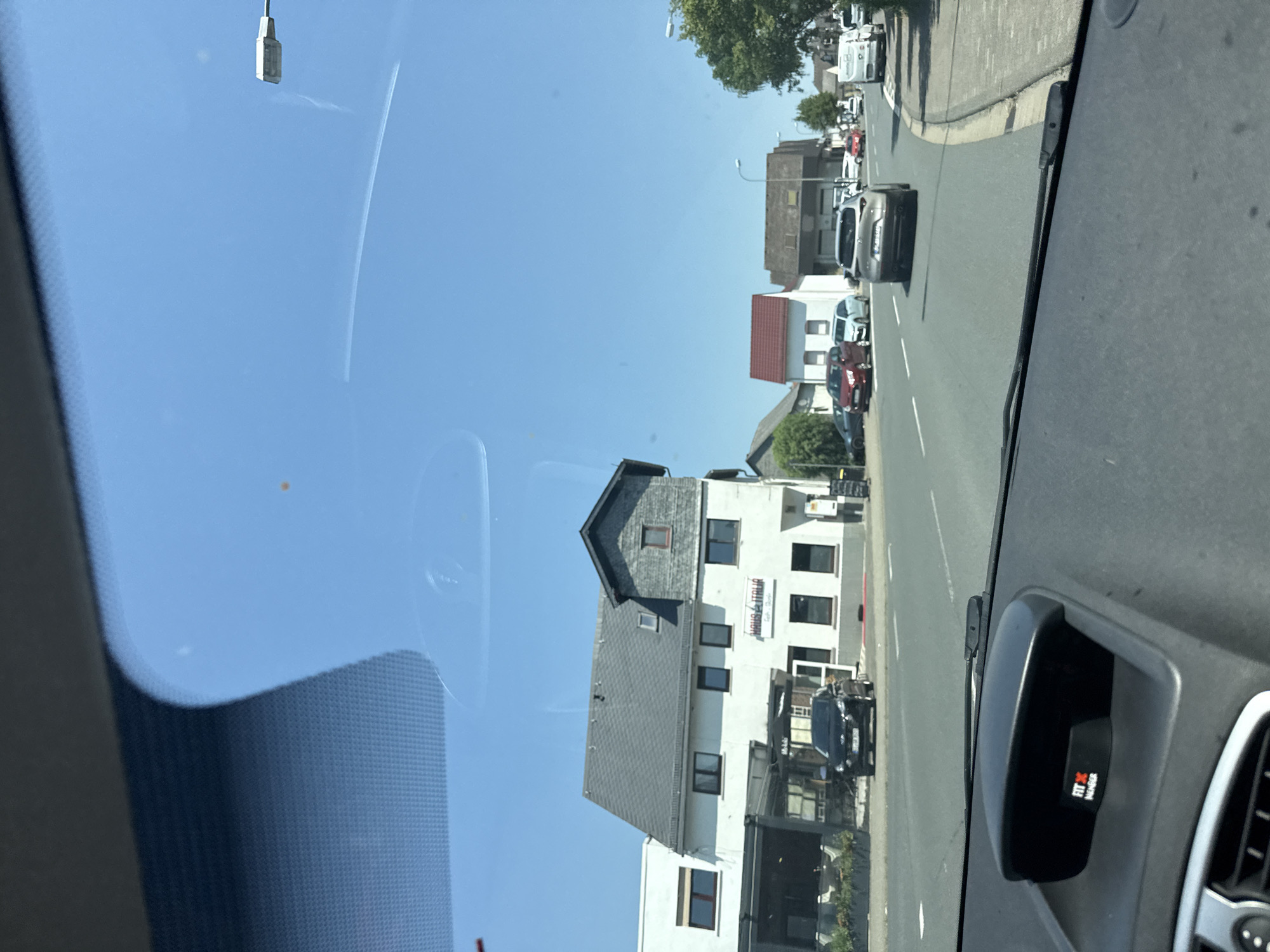 The image shows a street scene in Nauborn, Germany, as viewed from inside a vehicle. The sky is clear and blue. On the left side of the street is a building with a gray roof, featuring windows and a sign that reads "HAUS ITALIA." A black car is parked in front of this building. Further down the street, other buildings and cars are visible. The road has lane markings. A street lamp is visible in the distance. The foreground shows the interior of the vehicle, including part of the dashboard and a device with a "FIT X MEMBER" logo. The overall impression is a daytime view of a residential or mixed-use street.