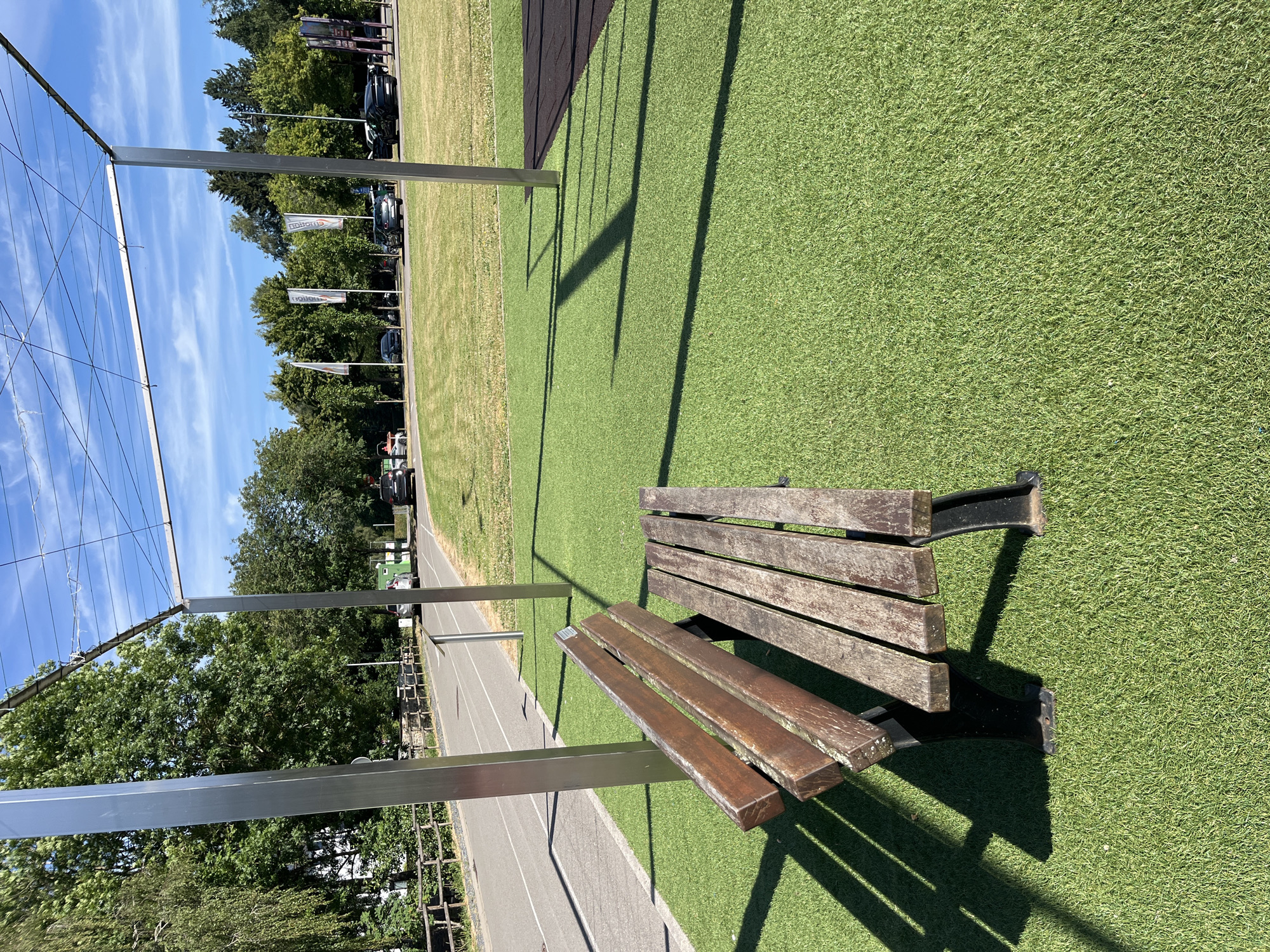 In the city of Saint-Vith, Belgium, a wooden park bench with black metal legs sits on artificial turf in front of a path bordering a grassy field. The bench is positioned under a modern metal pergola structure with thin wires stretched across its top. In the background, trees line the perimeter of the field, and several cars are parked or driving on a road. The sky is bright blue with scattered white clouds. The scene is illuminated by natural daylight, casting distinct shadows from the pergola onto the turf. There are no people present in the image.