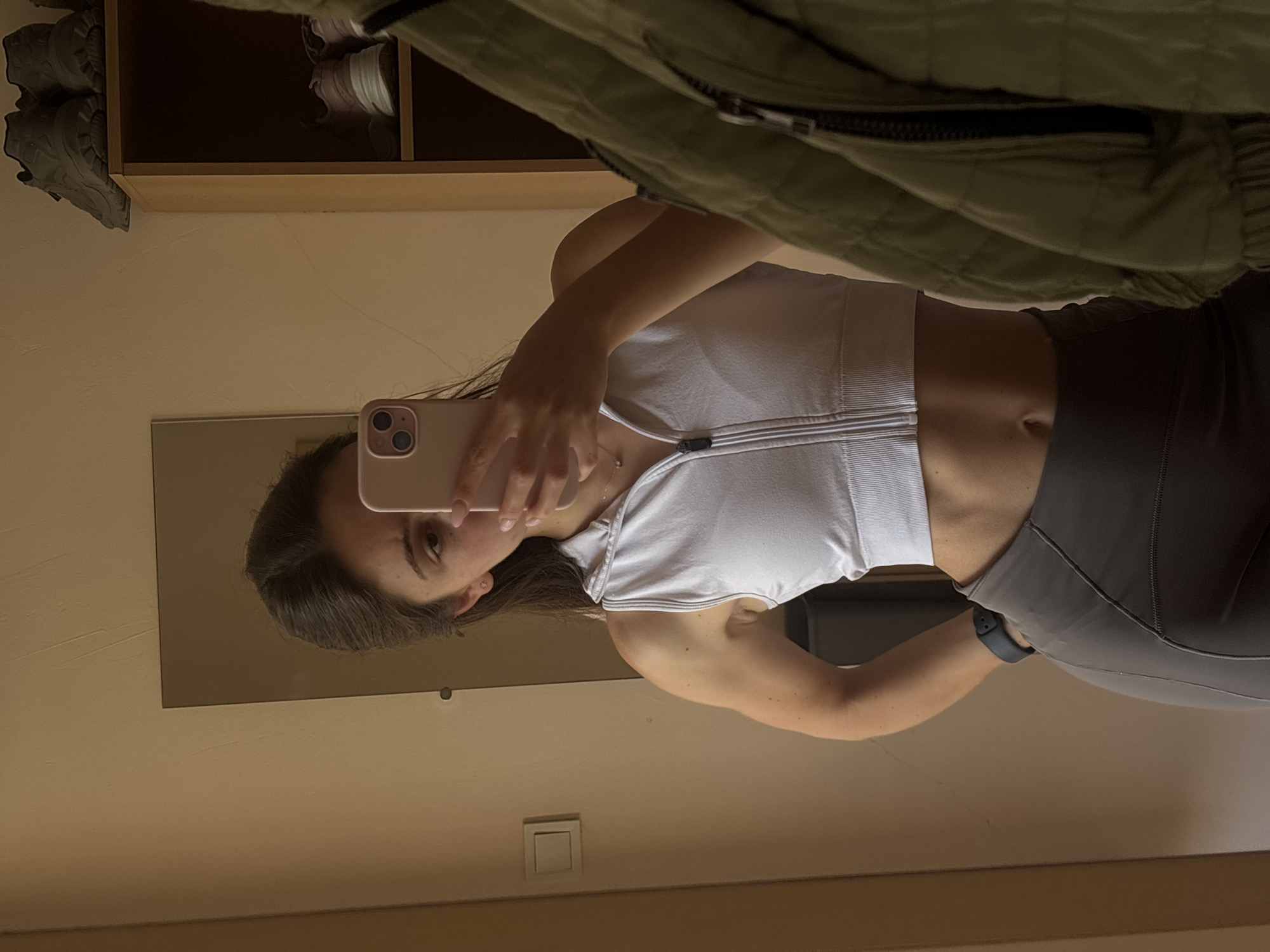 A young woman is taking a mirror selfie in what appears to be a room. She is wearing a white sports bra with a zipper down the front and grey athletic leggings. She has her hair down, and is holding a light pink smartphone in her right hand to take the picture. A green quilted jacket is draped over her left shoulder. In the reflection of the mirror, part of a closet with shelves and shoes can be seen. The room appears to have beige walls and a light wooden shelf. There is no visible text. The lighting suggests it is daytime, and the indoor setting provides no weather cues. The woman's expression is neutral. The context provided indicates the location is Varna, Bulgaria.