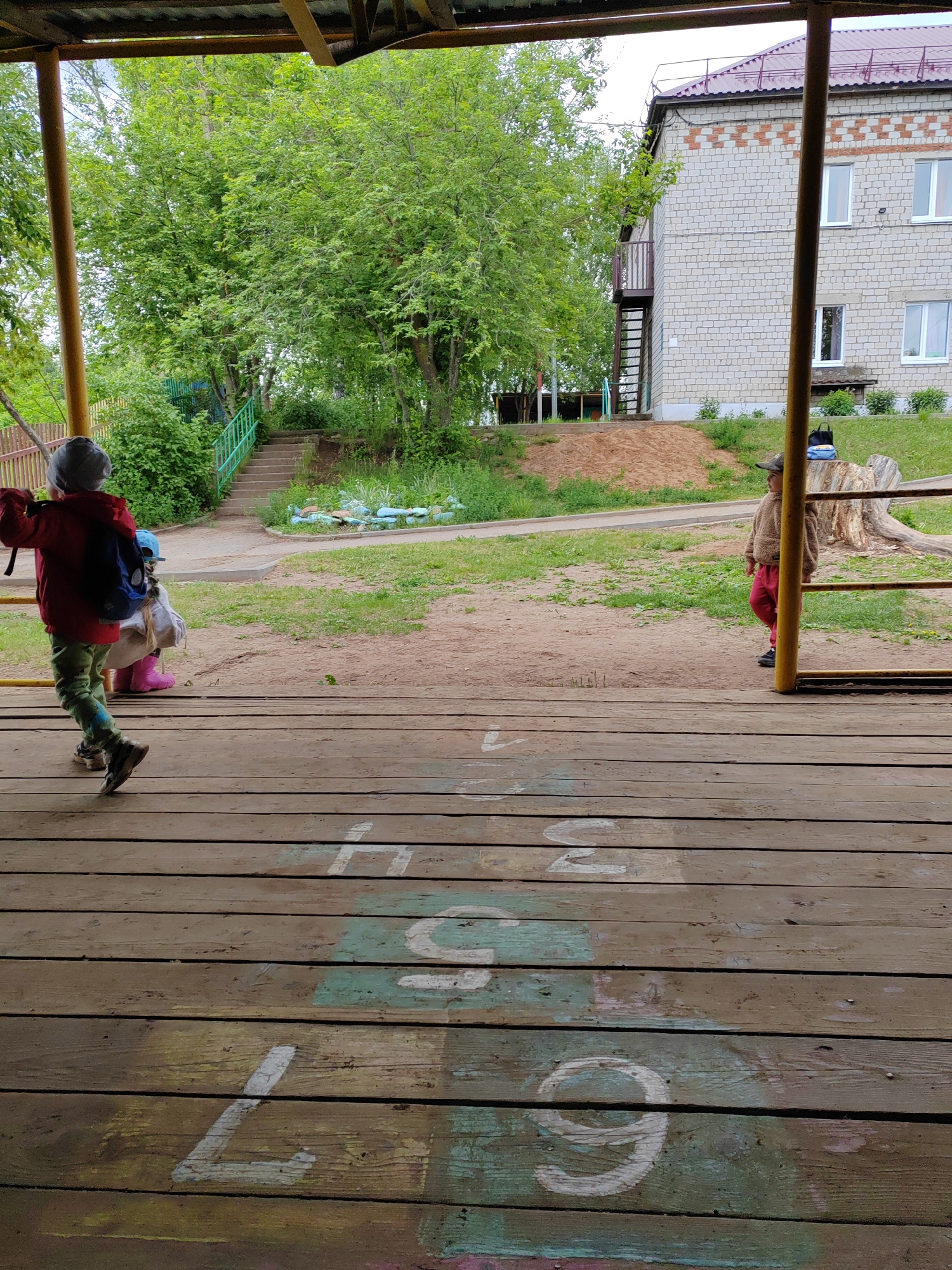 The image captures an outdoor scene, likely a playground or a yard of a facility, viewed from under a covered structure with a wooden floor. The foreground is dominated by the weathered wooden planks of a platform, on which several numbers are painted in white and faded colors, including "4", "5", "6", "7", and "9".
Two children are prominent, with a third partially visible. On the left, a child wearing a red jacket, green camouflage pants, a grey beanie, and a blue backpack is in motion, appearing to walk or swing, framed by a yellow metal support pole. Behind this child, another child is partially obscured but distinctive for wearing bright pink boots. On the right side of the frame, another child, dressed in a brown fleece jacket, red pants, and a baseball cap, stands beside another yellow metal support pole, looking towards the left.
The midground features a dirt path, patches of green grass, and some scattered objects, including a blue bag resting on a large tree stump. In the background, lush green trees and bushes create a natural backdrop. A concrete staircase with a green metal handrail ascends a grassy slope towards more trees. To the right of the stairs, a two-story building with light-colored brick walls and a red roof is visible. The building has several white-framed windows and an external metal staircase on its left side. A distinctive pattern of reddish-brown bricks decorates the upper part of the building's facade.
The weather appears overcast, suggesting a mild daytime, possibly late spring or early summer, given the vibrant greenery. The overall atmosphere is calm, with children engaged in unidentifiable play or movement.