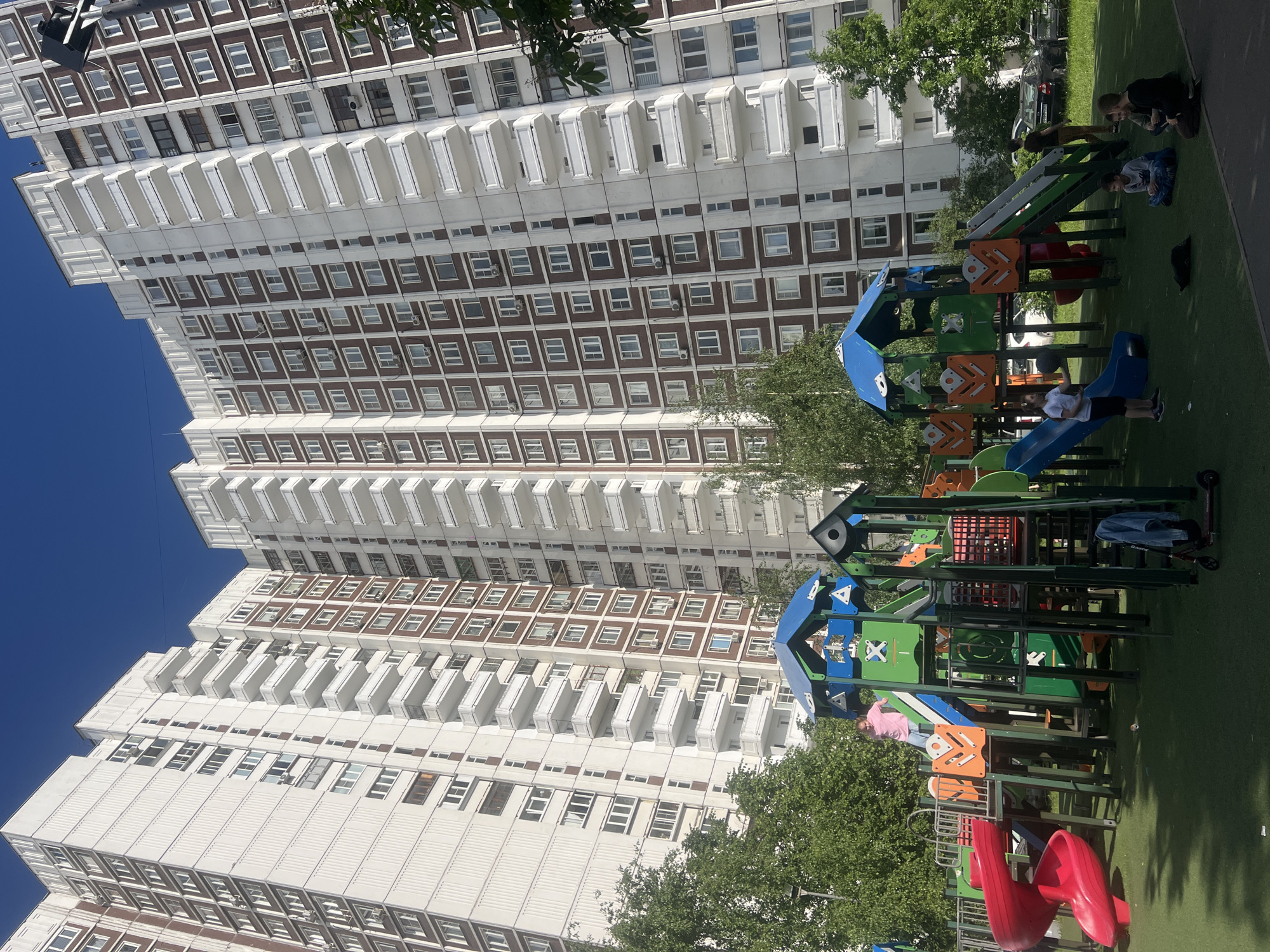 The image captures a vibrant outdoor scene on a clear, sunny day, set against the backdrop of a large residential building. In the background, a towering, multi-story apartment block dominates the left side of the frame. The building features a distinctive architectural pattern of alternating white horizontal bands with balconies and rows of brown rectangular sections containing numerous windows. Many of these windows have small air conditioning units.
In the foreground and to the right, a modern, colorful children's playground structure is the focal point. It boasts elements in bright blue, green, orange, and red, including multiple slides (a blue one and red ones), platforms, and climbing areas. Several children are actively playing on the structure; one child in a white t-shirt is seen on the blue slide, another in a light blue top sits on a green platform, and a child in a pink top is on an orange platform near the red slides. A small black and red scooter is parked near the playground.
On the artificial green turf that covers the playground area, a few adults are present, seemingly supervising the children. Two adults and one child are gathered towards the back right of the playground. Trees with green foliage are scattered around the area, providing some shade, and several parked cars are visible behind the trees in the mid-ground. The sky above is a clear, deep blue, indicating bright daylight. The overall atmosphere is one of leisure and activity in an urban residential setting, consistent with Moscow, Russia. No discernible text is visible in the image.