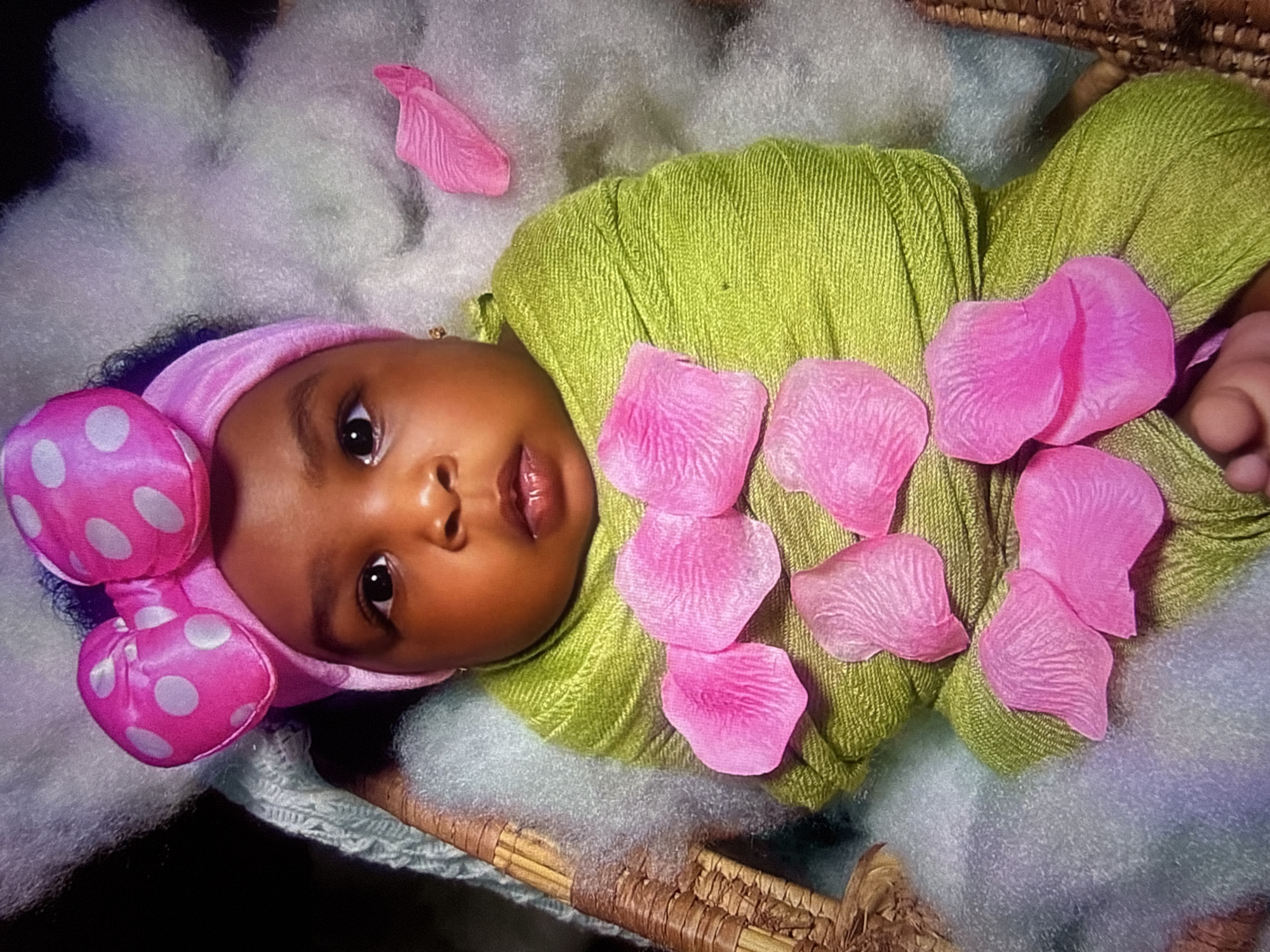 A baby is pictured wrapped in a lime green fabric, adorned with pink rose petals. The baby wears a pink headband with a large pink bow featuring white polka dots. The baby is lying in a woven basket filled with fluffy white material. The background is dark. The image does not provide any information about its location in Abuja, Nigeria.