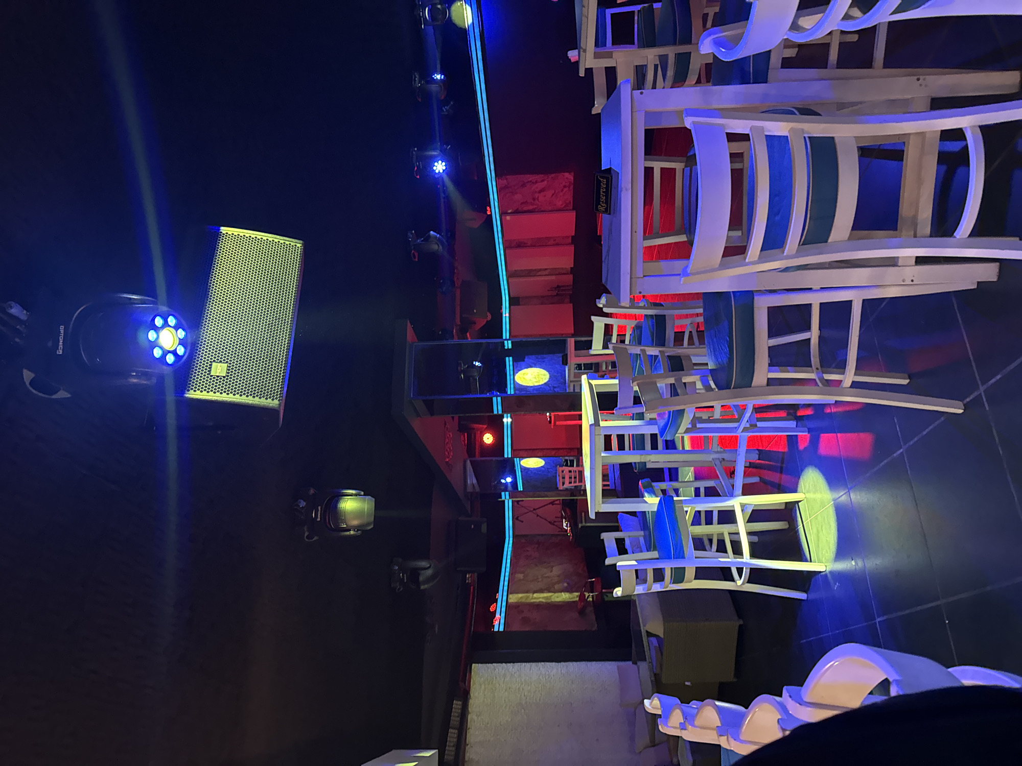 The image depicts an indoor scene, likely a bar or restaurant in Plovdiv, Bulgaria, due to the presence of a 'Reserved' sign written in Cyrillic. The location is lit with neon and LED lighting, casting blue, red, and yellow hues. There are stacks of white and blue outdoor chairs and tables, and a sound system. The image is rotated, with the right side appearing to be the 'top' of the scene.