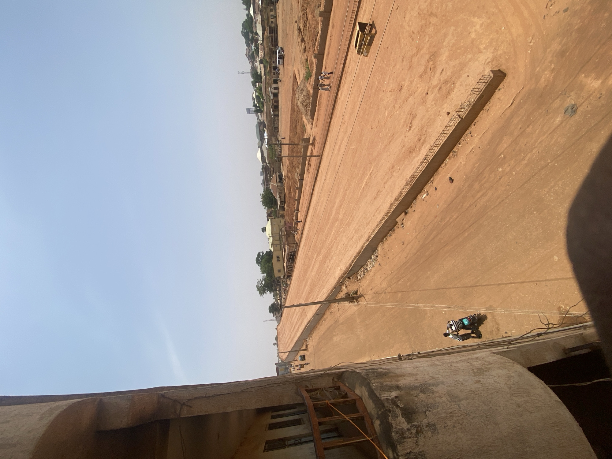 An elevated view looking down upon a wide, dirt road in Tudun Wada, Nigeria. The road is bordered by concrete structures and appears to be under construction. Several people are seen working along the road. Buildings and vegetation line one side of the road. A utility pole with wires is visible. In the foreground is part of the rooftop of a building. The sky is a clear light blue.